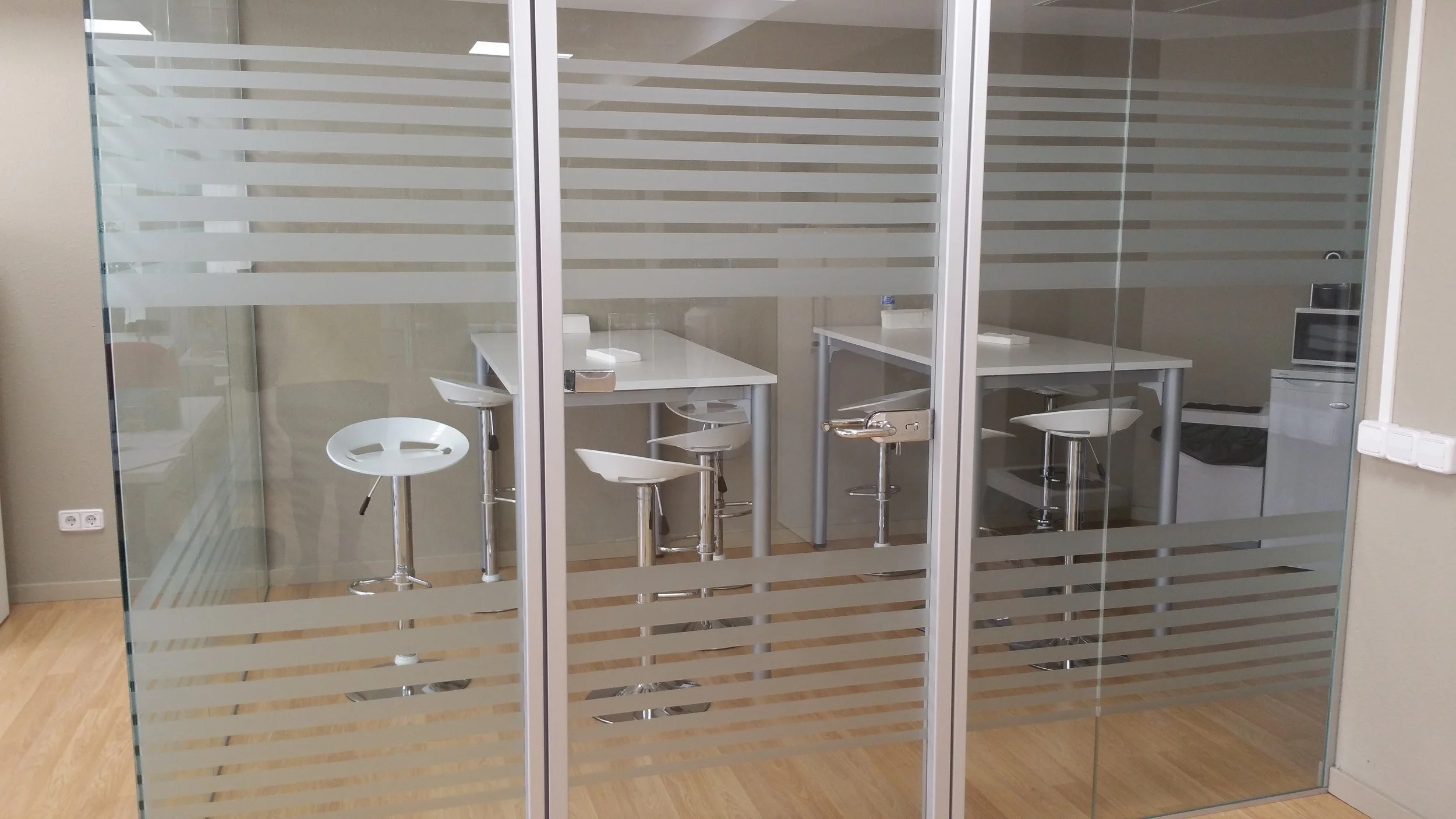 Una sala moderna con paredes de vidrio con franjas horizontales y sillas altas blancas con pedestal de metal, una mesa blanca y algunos objetos visibles al fondo.