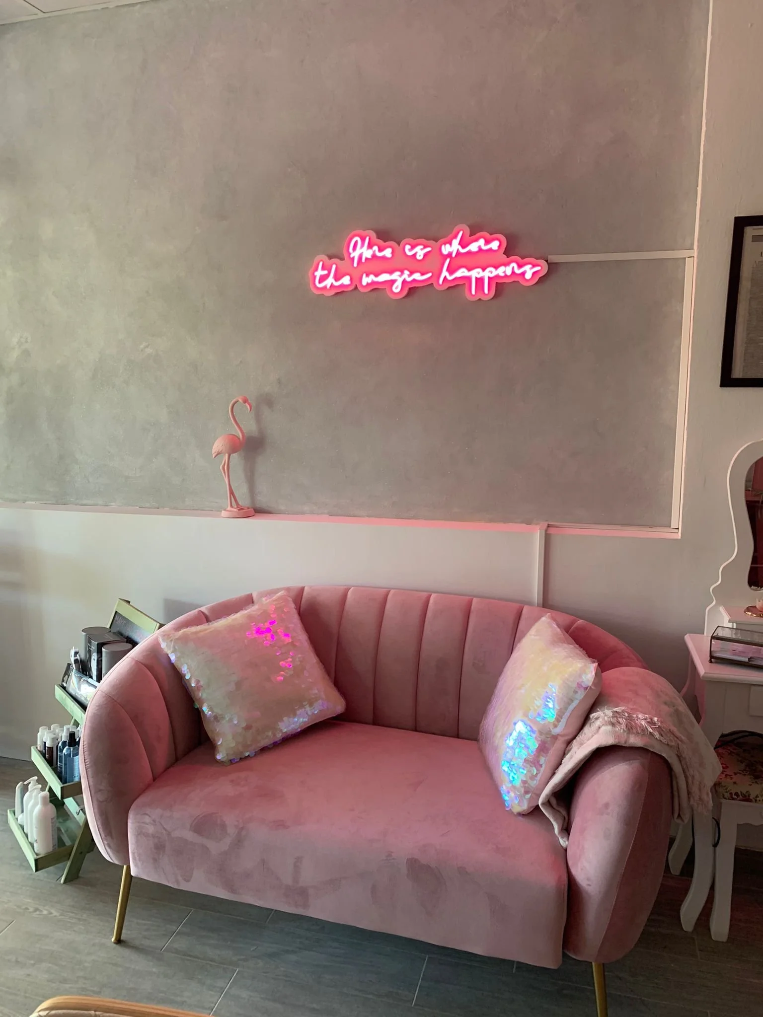 Sala decorada en tonos rosa con un sofá curvo de terciopelo, con cojines de purpurina y una manta. En la pared una cita en neón rosa que dice 'Here is where the magic happens' y una figura de flamenco rosa en una repisa.