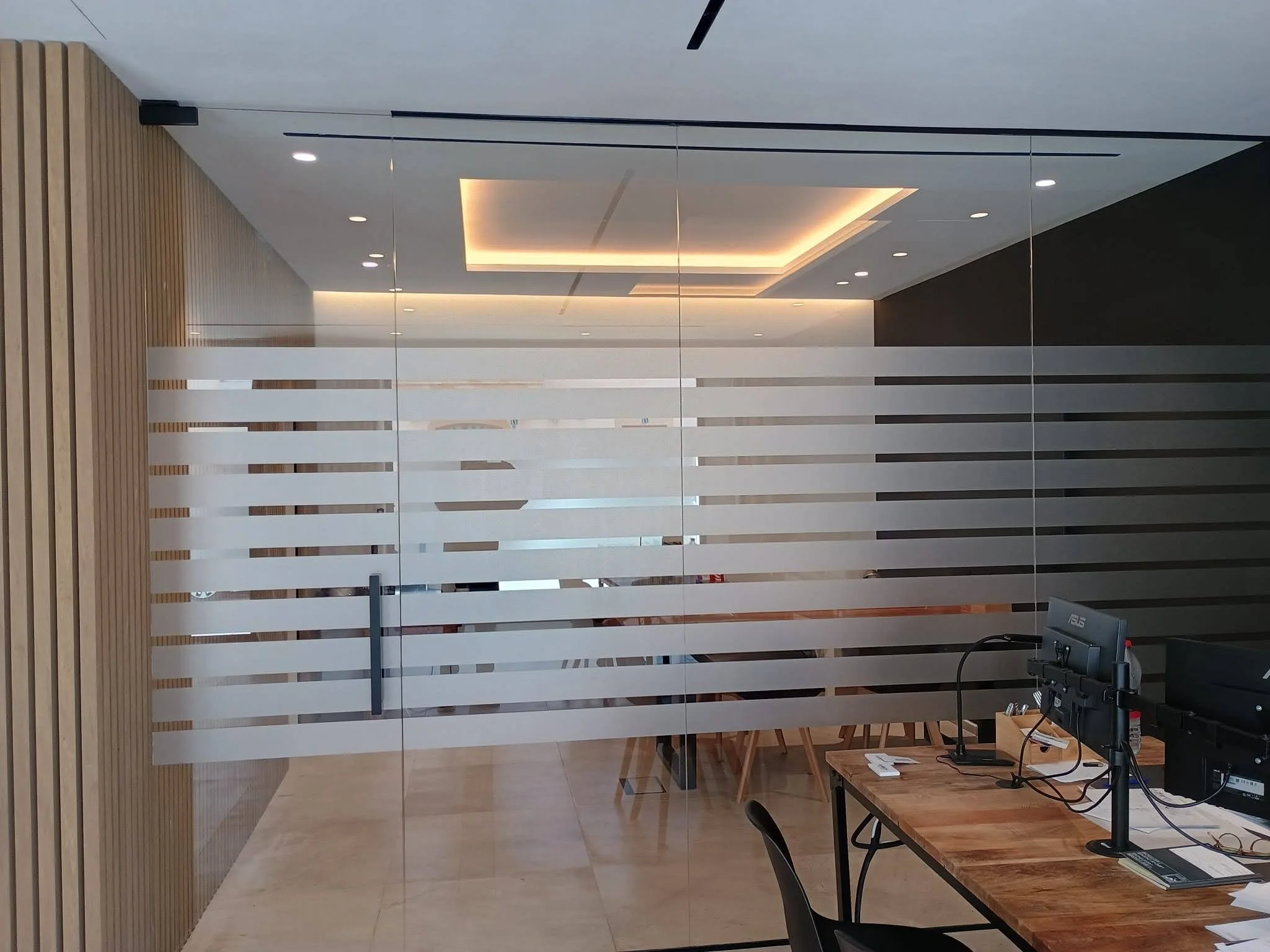 Interior de oficina moderna con paredes de madera y división de vidrio con franjas horizontales blancas, mesa de trabajo con monitor y silla.