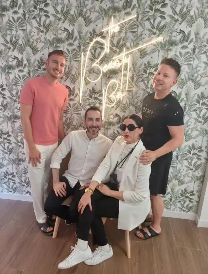Cinco personas posando en una sala con paredes decoradas con papel tapiz de hojas y una señal luminosa que dice 'The Beauty Bar'.