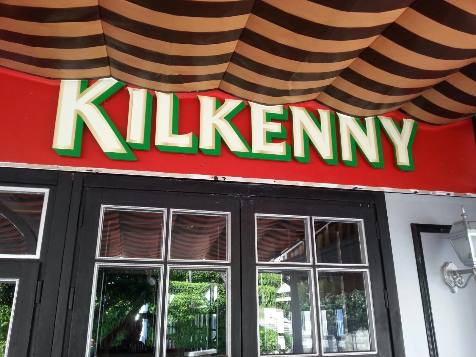 Cartel con letras grandes que dice 'KILKENNY' en la fachada de un establecimiento, con ventanas y puertas negras y un toldo con franjas marrón oscuro y beige.
