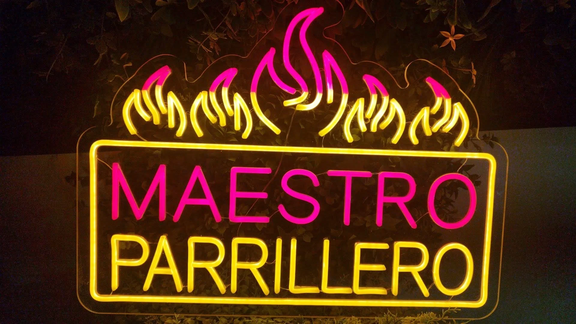 Cartel de neón con letras rosas y amarillas que dice 'Maestro Parrillero' con llamas en colores rosa y amarillo en la parte superior