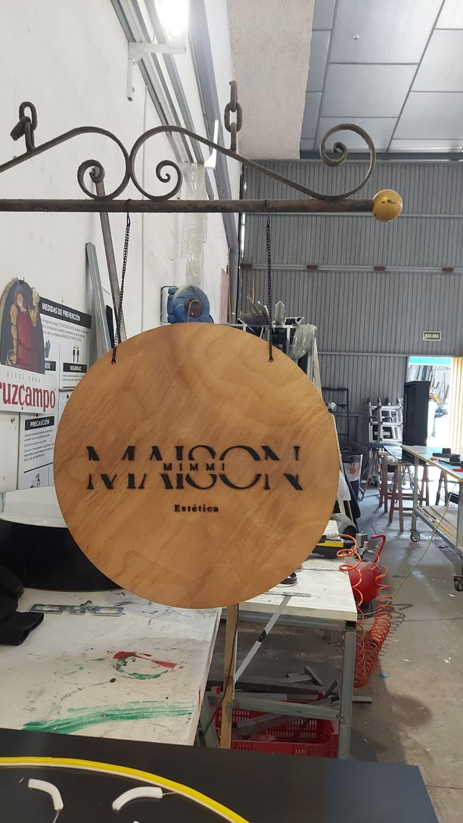 Pieza de madera con letras cortadas que dicen 'MALCON Estética', colgada en un taller de carpintería o artesanía.