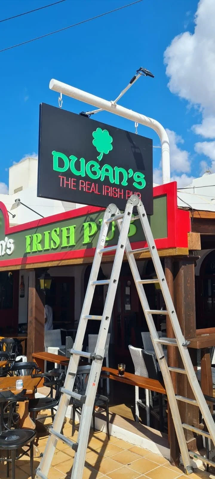 Cartel en un pub irlandés llamado Dugan's, con un letrero que dice "Dugan's, The Real Irish Pub" y "Irish Pub" en la fachada, en una zona exterior con mesas y sillas, y una escalera de aluminio en primer plano.