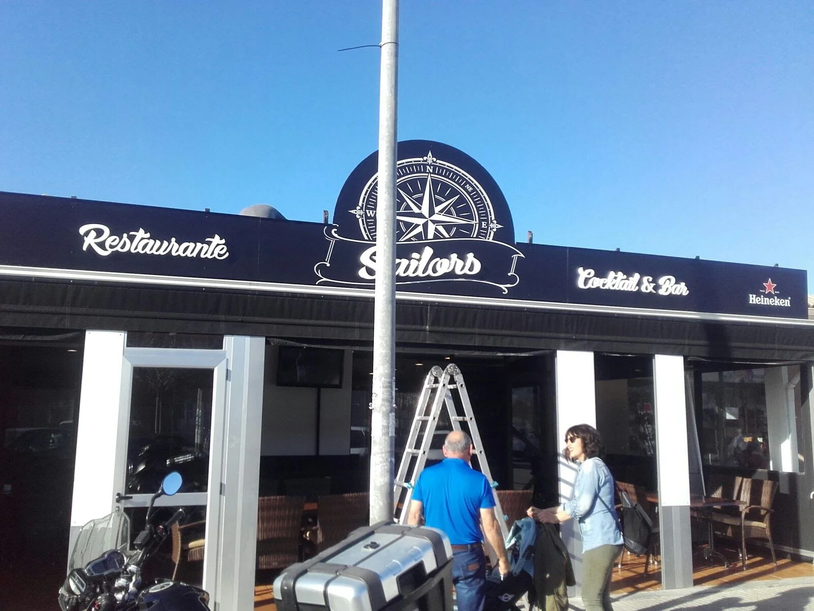 Exterior de un restaurante llamado Sailors, que ofrece cócteles y tiene la marca Heineken, con personas entrando, un coche y una motocicleta en la acera, en un día soleado con cielo despejado.