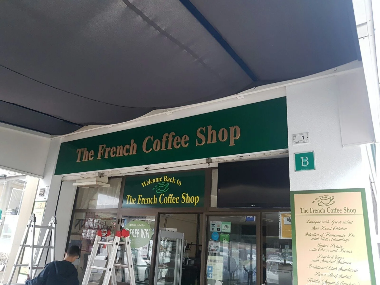 Entrada de una cafetería llamada 'The French Coffee Shop' con un cartel que indica 'Welcome Back to The French Coffee Shop'.