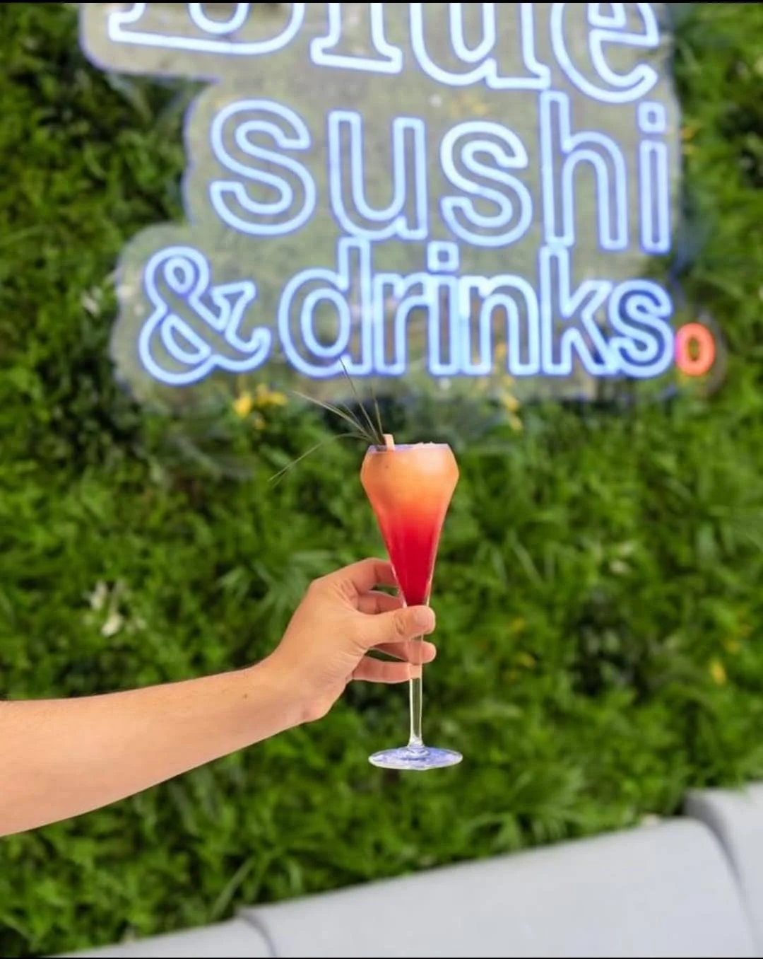 Vaso con cóctel rosado y rojo en forma de copa de champán, con fondo de pared con letrero de neón que dice 'Blue sushi & drinks' y vegetación verde.