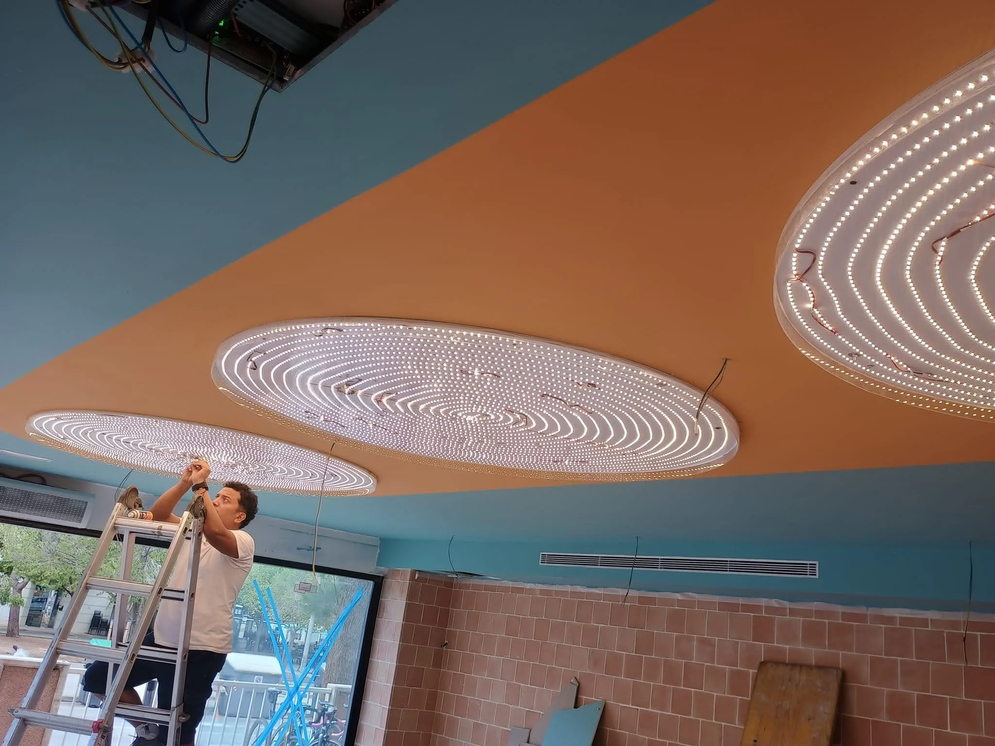 Un hombre de pie en una escalera ajustando la iluminación del techo en un interior con paredes de ladrillo y un techo decorativo con luces LED. El fondo muestra ventanas que permiten la vista del exterior.