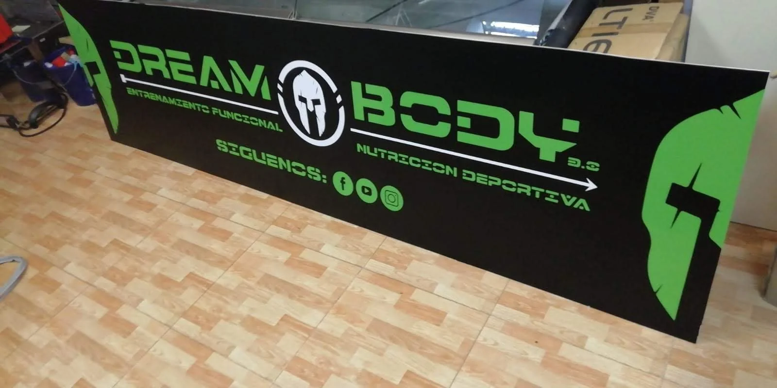 Cartel negro y verde con logotipo de un casco griego y texto que promociona un gimnasio llamado 'Dream Body' y las redes sociales Facebook, YouTube e Instagram.