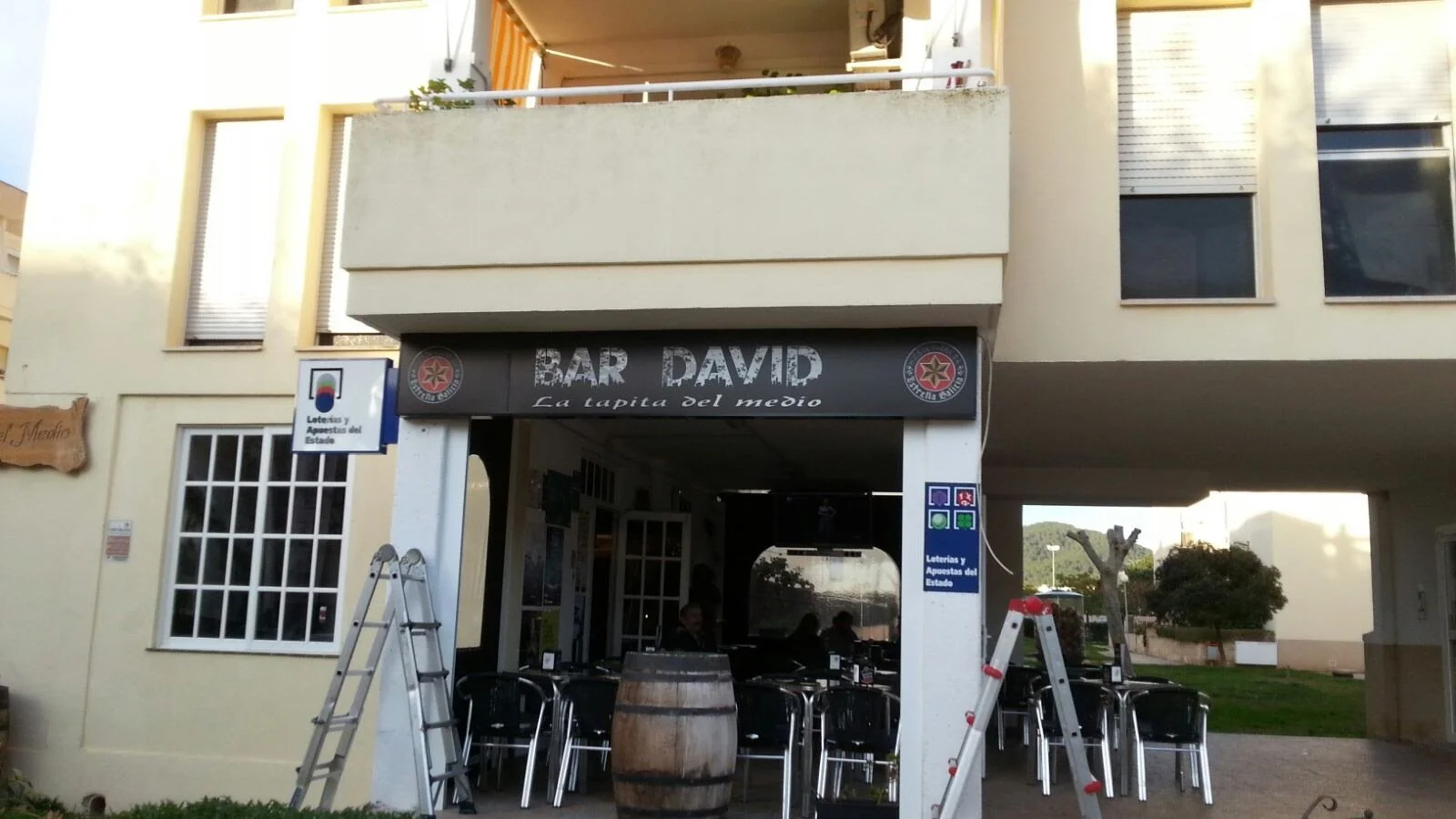 Vista frontal de un restaurante llamado Bar David con mesas y sillas, decorado con un barril de madera y mesitas exteriores, en un ambiente urbano.