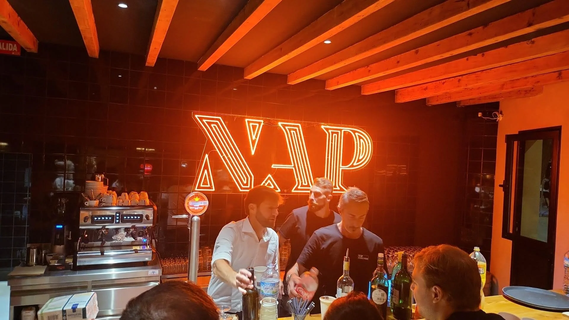Interior de un restaurante con un cartel de neón que dice 'XAP' en la pared negra, y personas sirviendo bebidas y atendiendo en la barra.