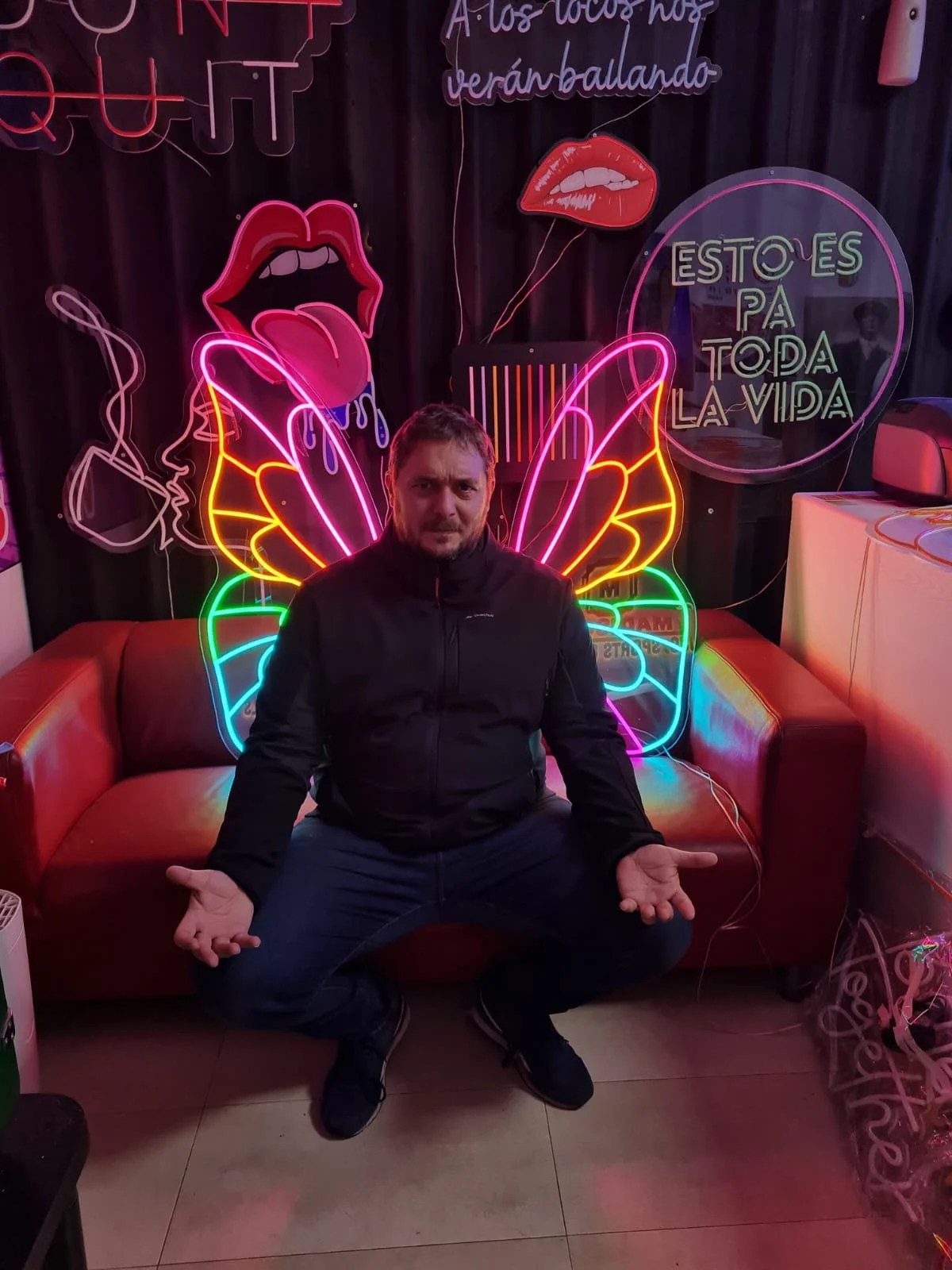Hombre sentado en un sillón con fondo de luces de neón coloridas, incluyendo labios y labios con lengua, y cartel que dice "Esto es para toda la vida".