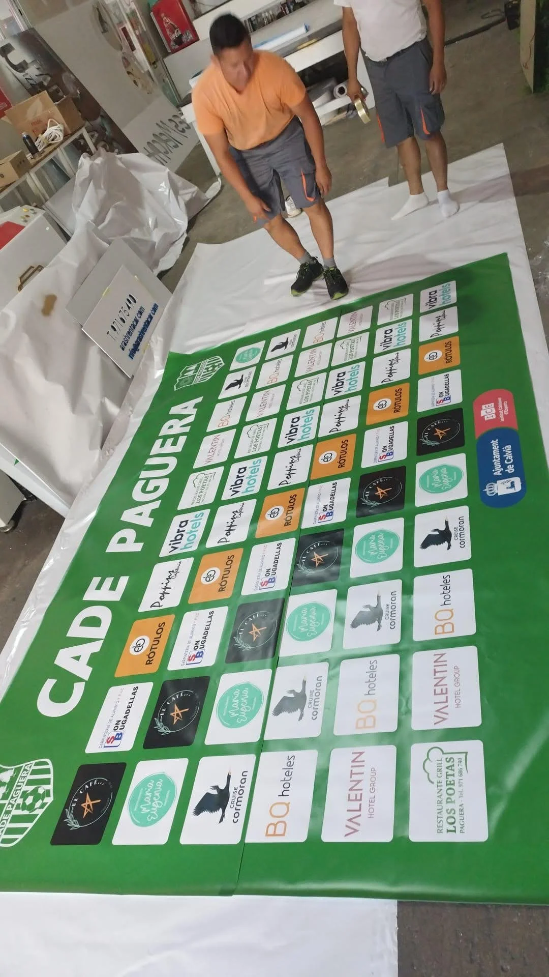 Una mesa con una cartulina grande de color verde que parece ser una especie de rifa o sorteo, con muchas casillas y logotipos de diferentes hoteles y empresas en ellas, y dos personas de pie observando la cartulina en un ambiente de almacén o taller.