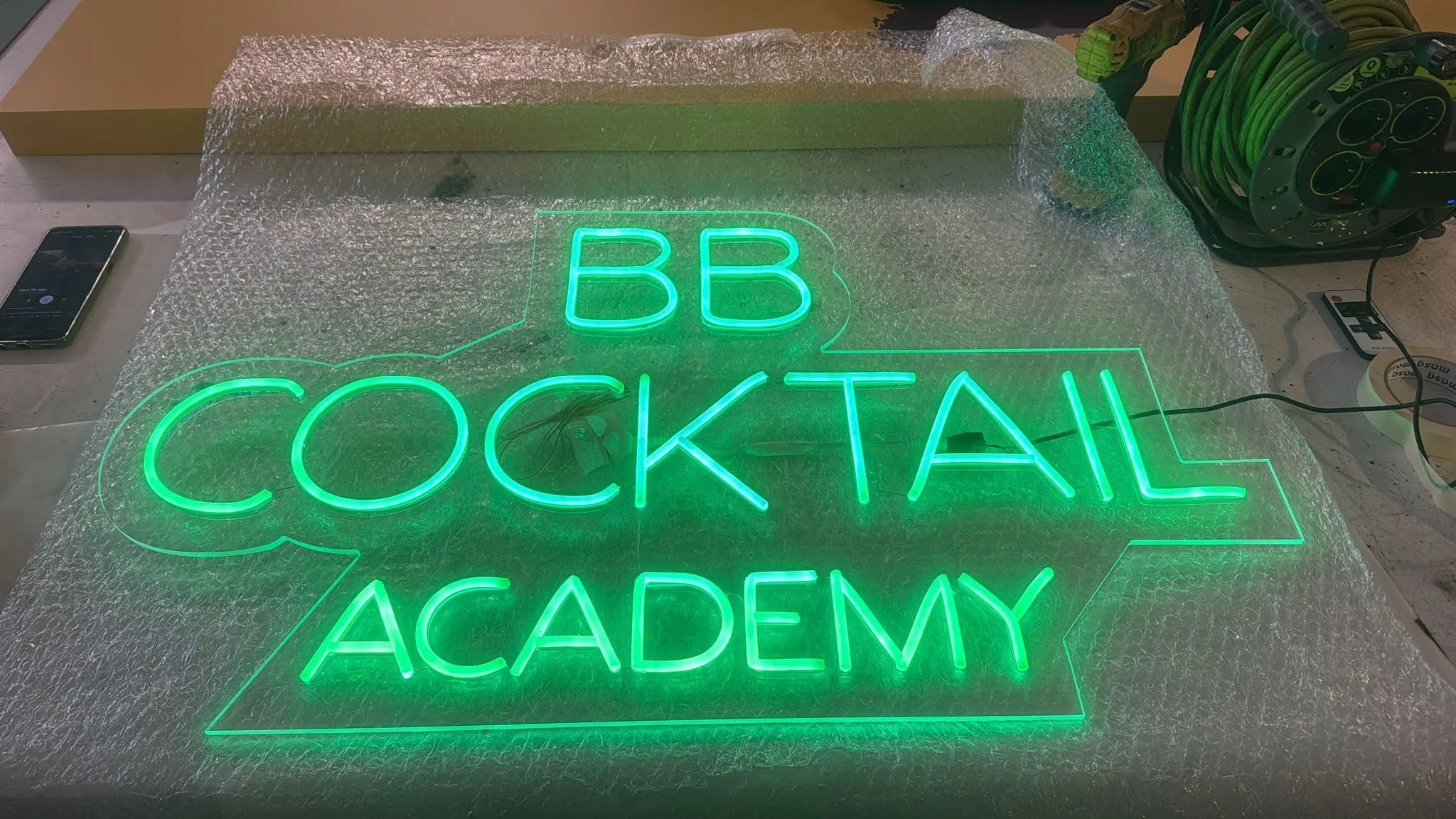 Letrero de neón verde que dice 'BB COCKTAIL ACADEMY' sobre una superficie de burbujas plásticas.