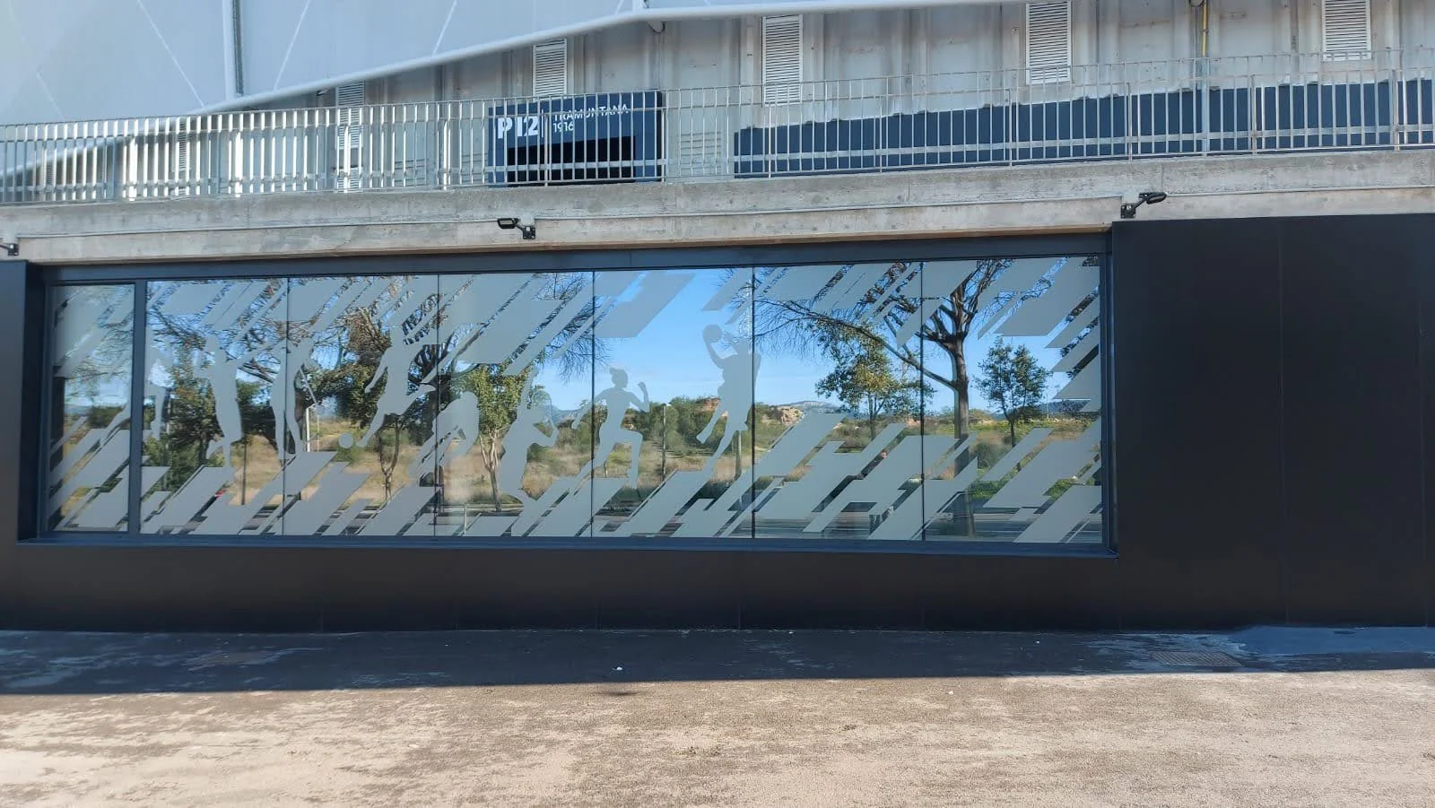 Ventana de vidrio con siluetas de personas corriendo y caminando, decoradas con franjas diagonales blancas y grises, en un edificio con paredes negras y una vista de árboles y cielo reflejada en el vidrio.