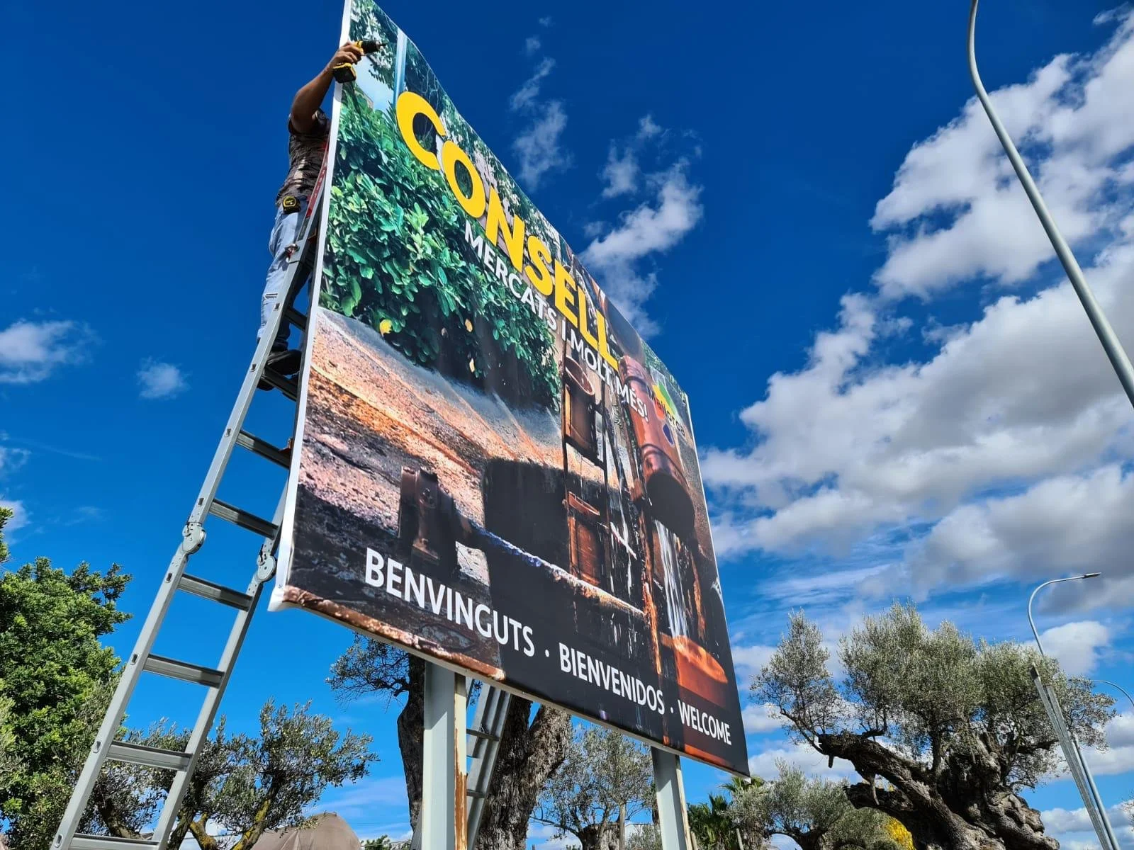Un cartel grande que da la bienvenida en varios idiomas y está siendo instalado con una escalera, en un día soleado con cielo azul y algunas nubes.