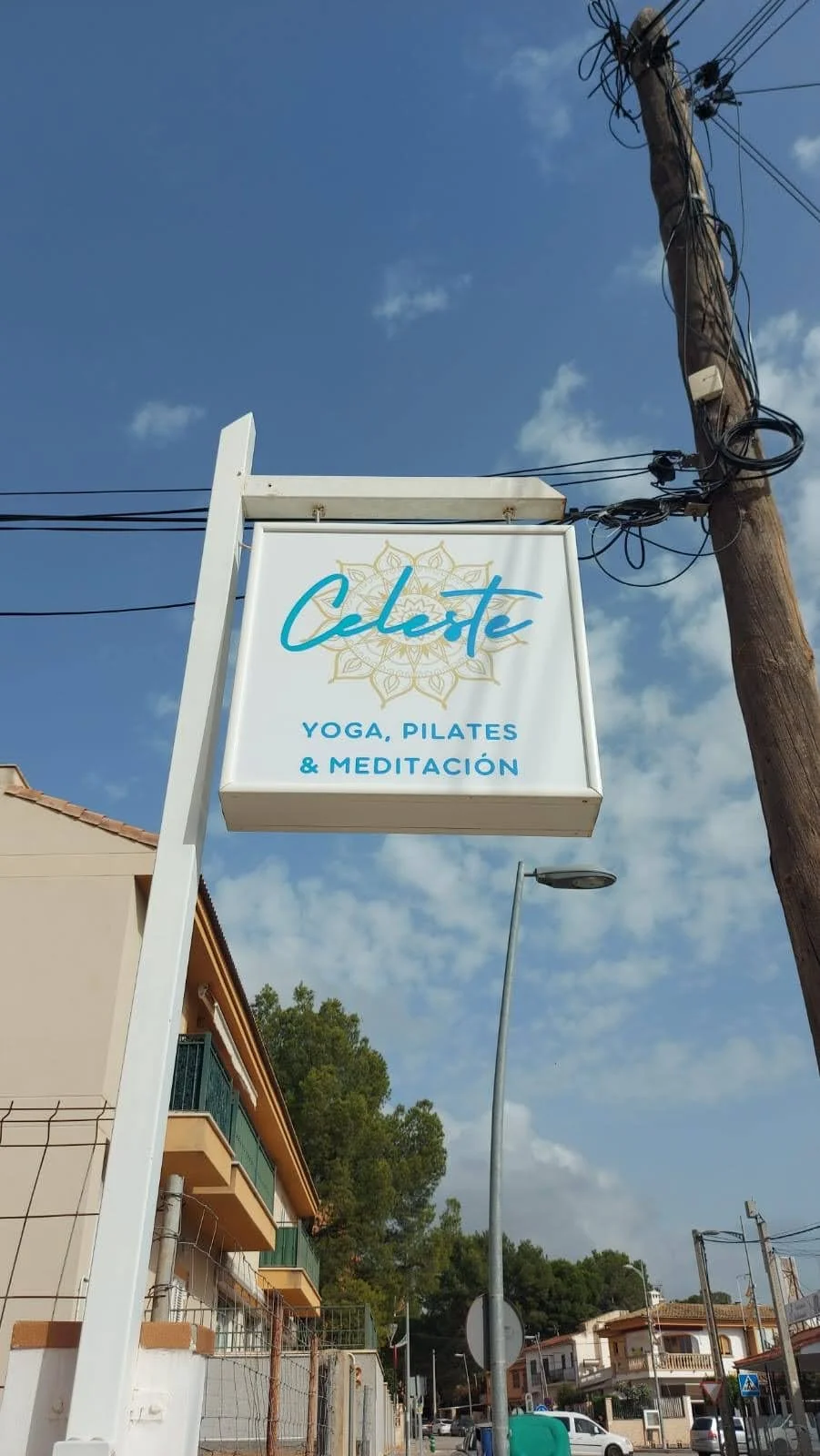 Cartel de un centro de yoga llamado 'Celeete' que ofrece clases de yoga, pilates y meditación, en un ambiente exterior con árboles y casas alrededor.