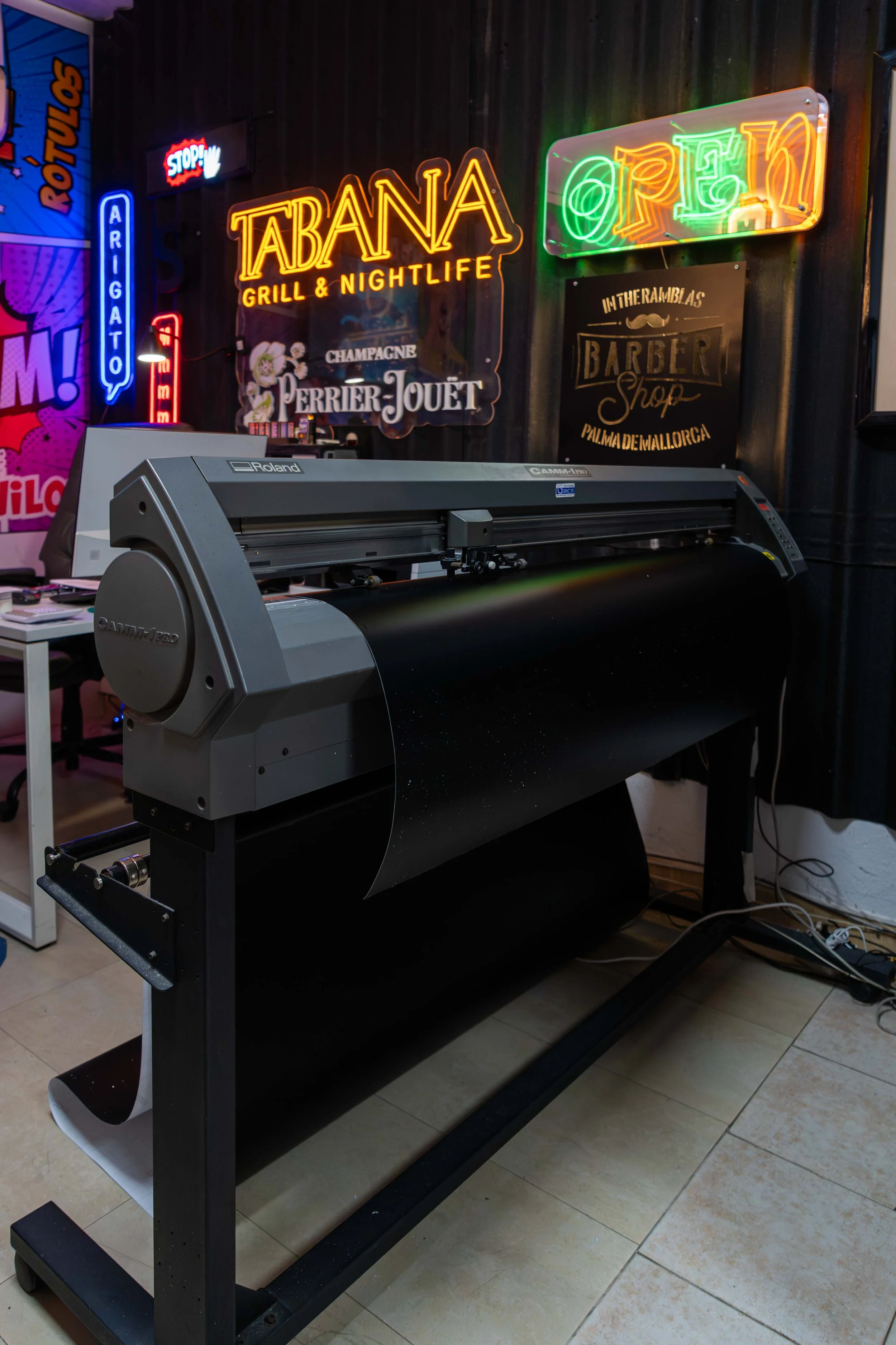 Impresora de corte de vinilo en un espacio con señales de neón en la pared que promocionan un lugar llamado Tabana, bar y área de noche, con otros anuncios de neon y un cartel de barbería en Mallorca.