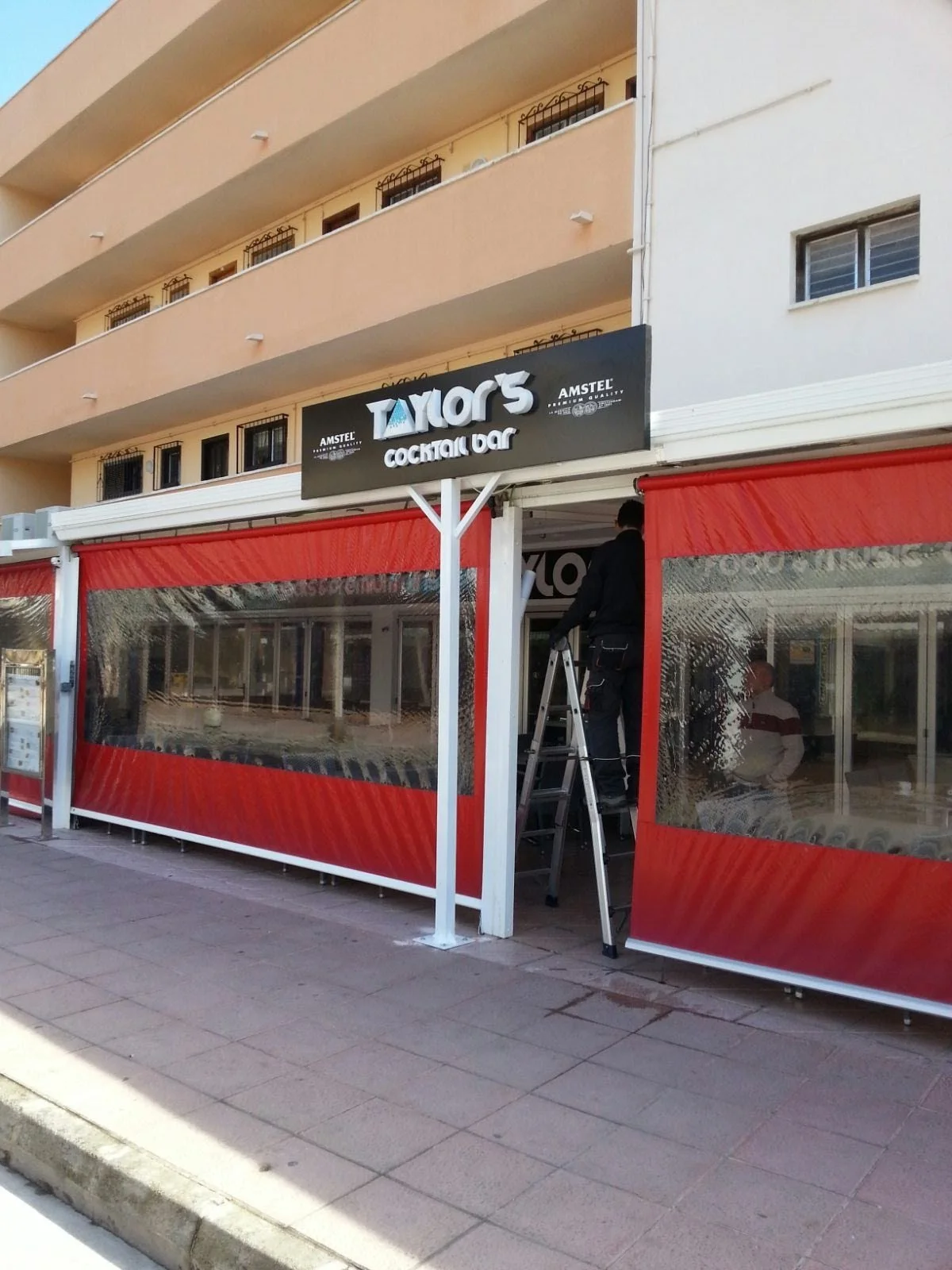 Exterior de bar llamado 'TAMOR'S Cocktail Bar' en construcción o reparación, con una persona en una escalera trabajando en la señal del negocio, con paredes con cortinas rojas y ventanas grandes con rejas.