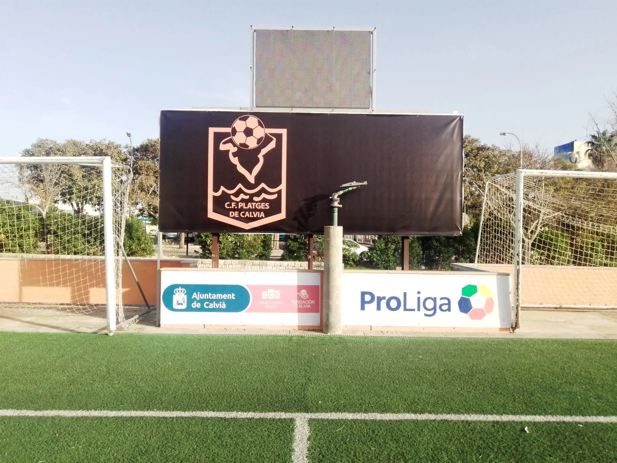 Cancha de fútbol con anuncios publicitarios y logos, césped artificial en primer plano, cielo despejado.