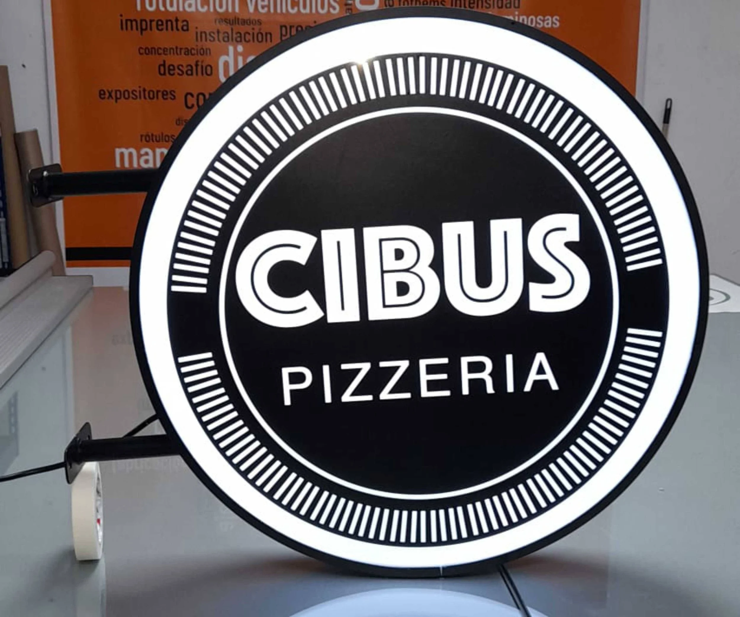 Reloj de neón circular en color negro y blanco con la palabra 'CIBUS' en el centro, y la palabra 'PIZZERIA' debajo, en un fondo oscuro.