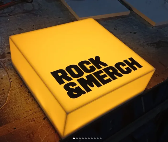 Lámpara cuadrada amarilla con letras negras que dice "ROCK & MERCH".