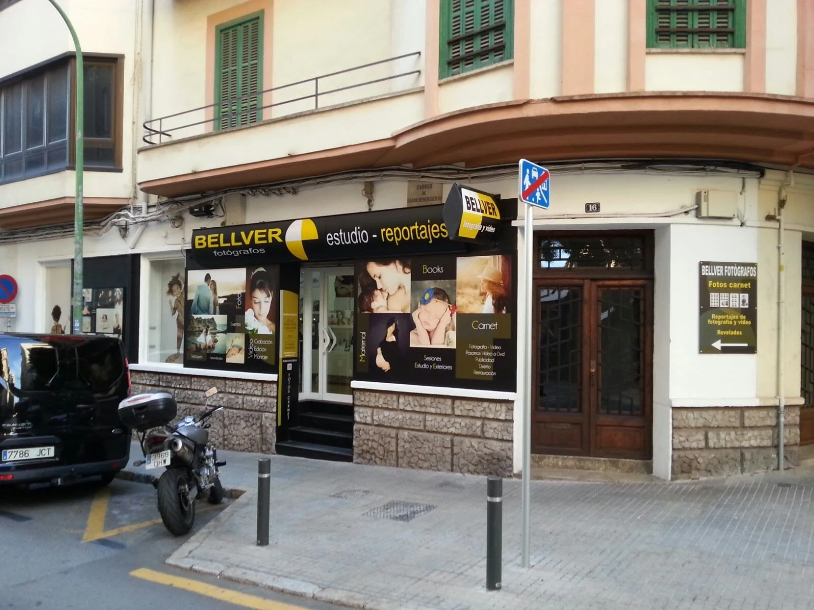 Fachada de un estudio de fotografía y reportajes llamado BELLVER, con ventanas verdes y cartelera publicitaria en la entrada, sobre la acera con motocicleta y coche negro estacionados enfrente.