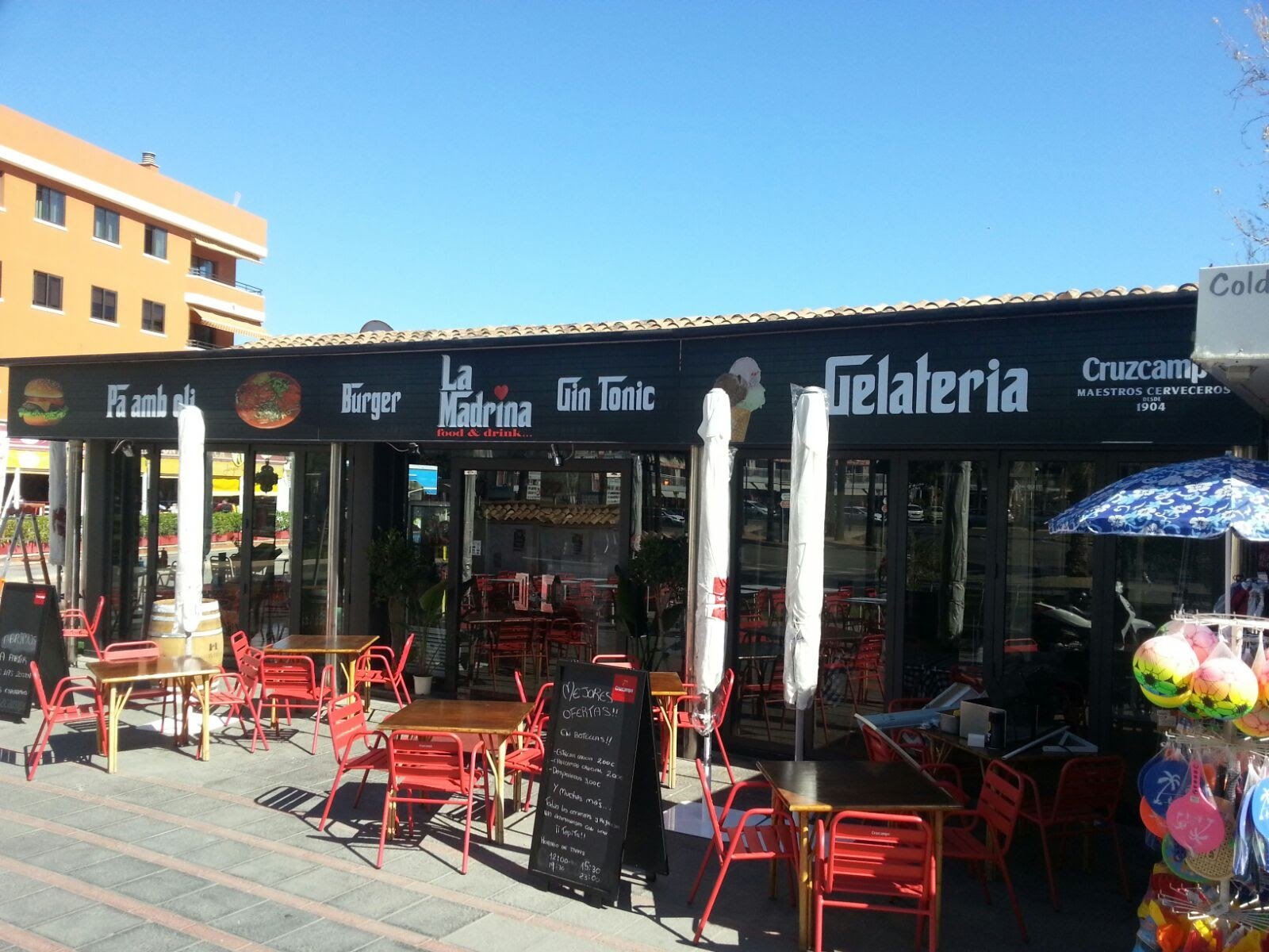 Fachada de una heladería con mesas y sillas afuera, cartel con ofertas y zona de venta de globos y otros artículos en la acera.