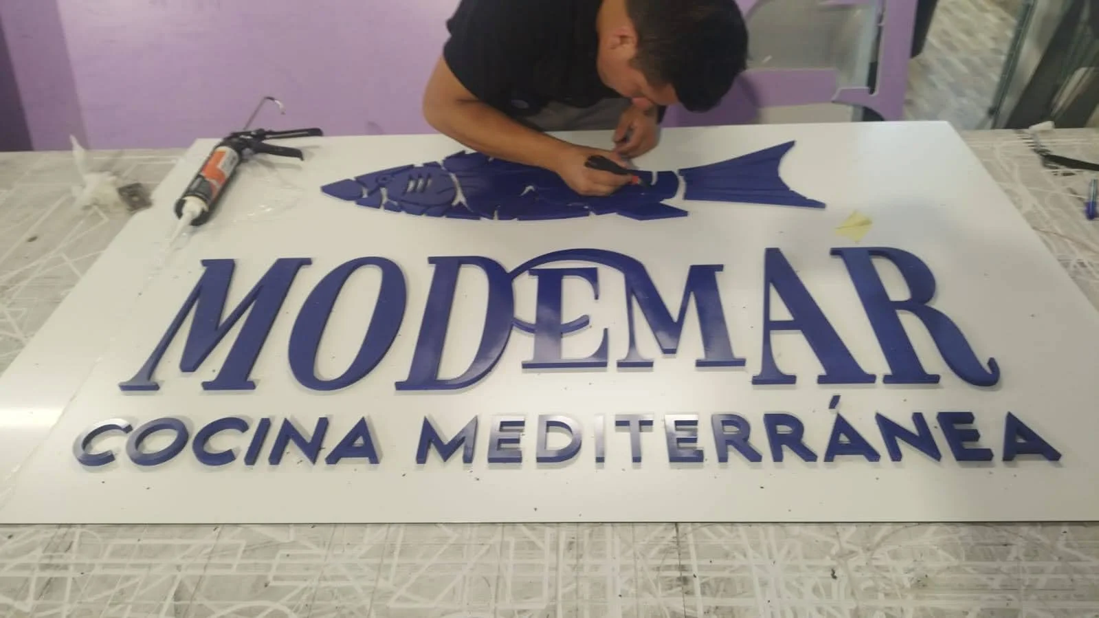 Persona ensamblando un letrero de la cocina mediterránea 'MODI MAR' con forma de pez en un taller.