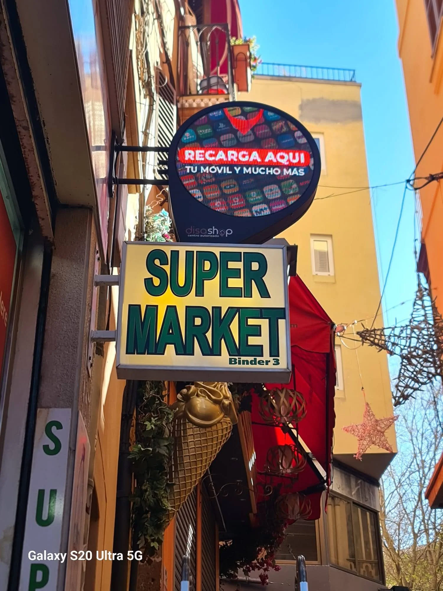 Cartel publicitario digital que dice: 'Recarga aquí tu móvil y mucho más'. Debajo hay un cartel que dice 'Super Market'. Estanterías con objetos decorativos y plantas, y un fondo de edificios y árboles con cielo despejado.