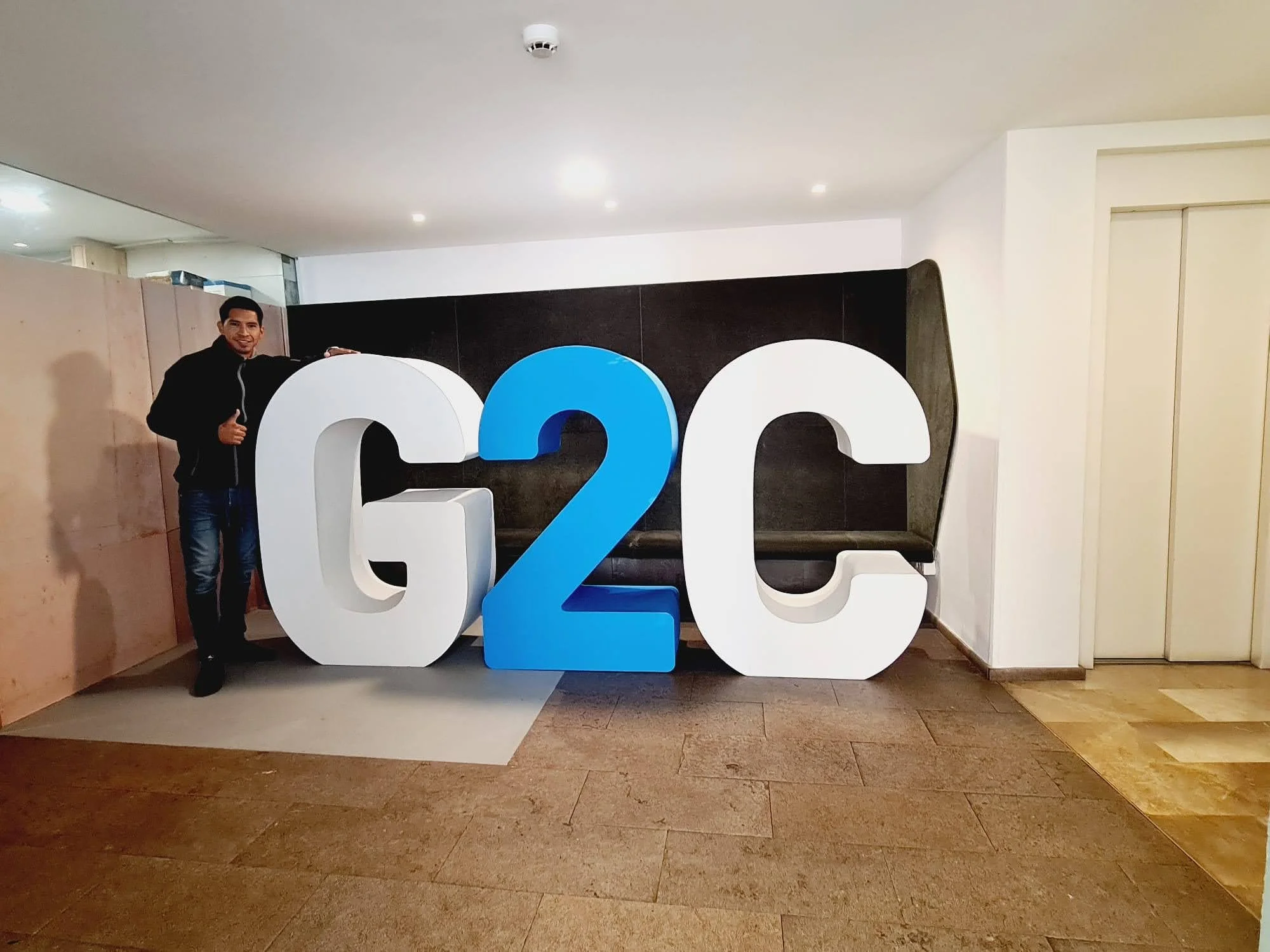 Hombre sonriendo, vestido con chaqueta negra, posando junto a una gran figura de letras blancas y azul que dicen '2022', en un espacio interior con paredes blancas y paredes de madera.