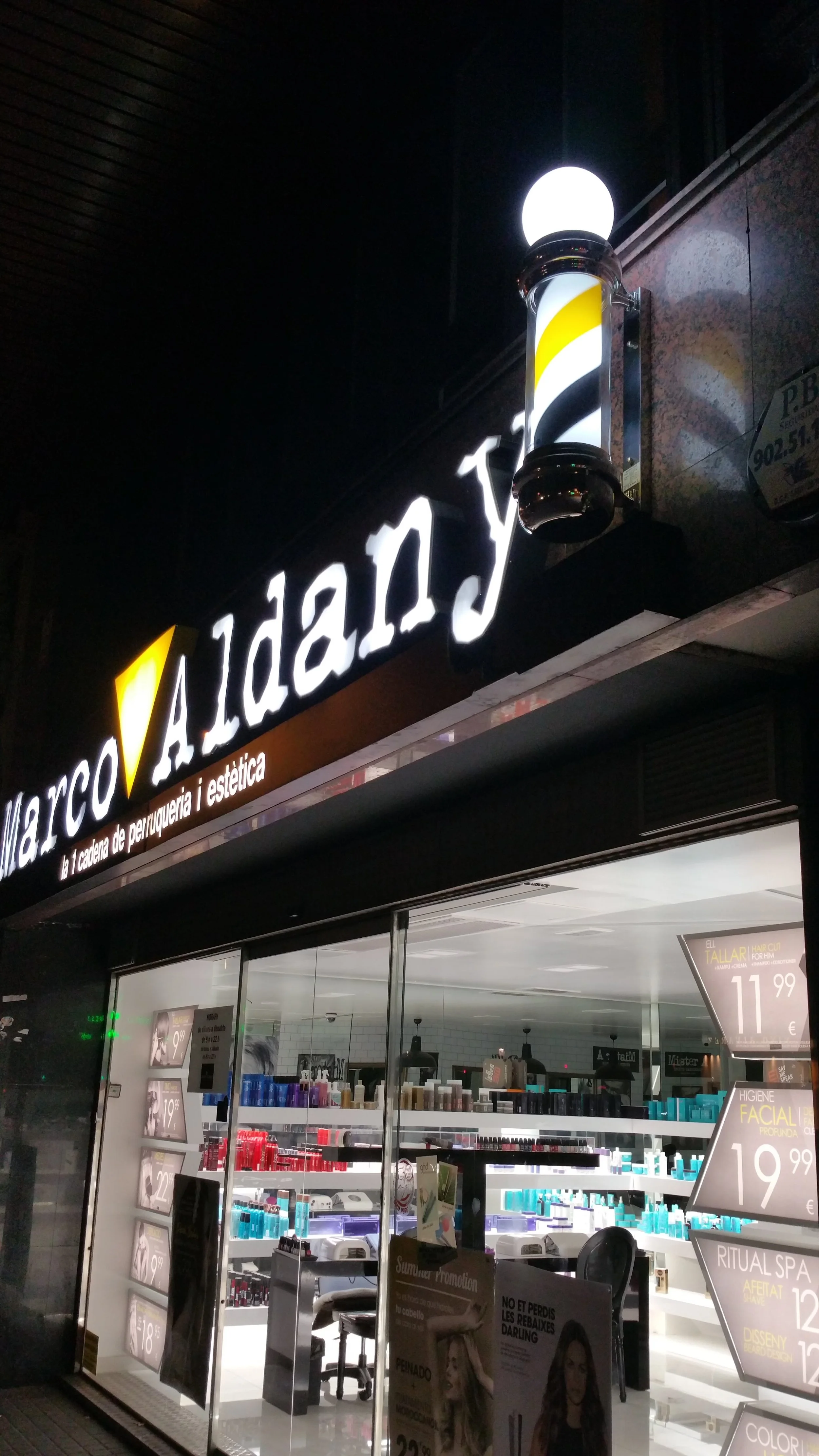 Vista nocturna de una peluquería y estética llamada 'Marco Amedy', con iluminación en el exterior y productos de belleza en el interior visible a través del escaparate, además de cartelera con promociones y precios.