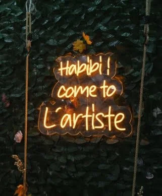 Cartel de neón que dice '¡Hable! Ven al artista', con fondo de follaje verde.