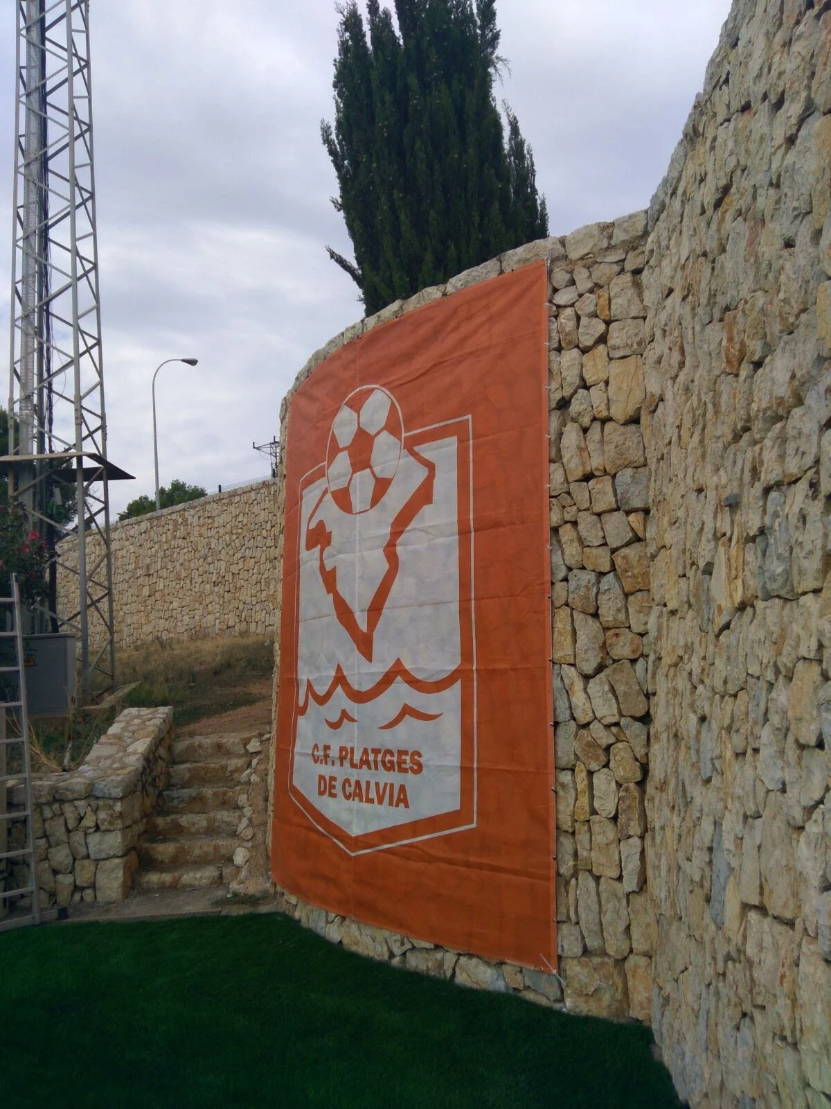 Una bandera naranja con el escudo del club de fútbol 'C.F. PLATGES DE CALVIA' colgado en una pared de piedra, en un área con césped artificial y árboles al fondo.