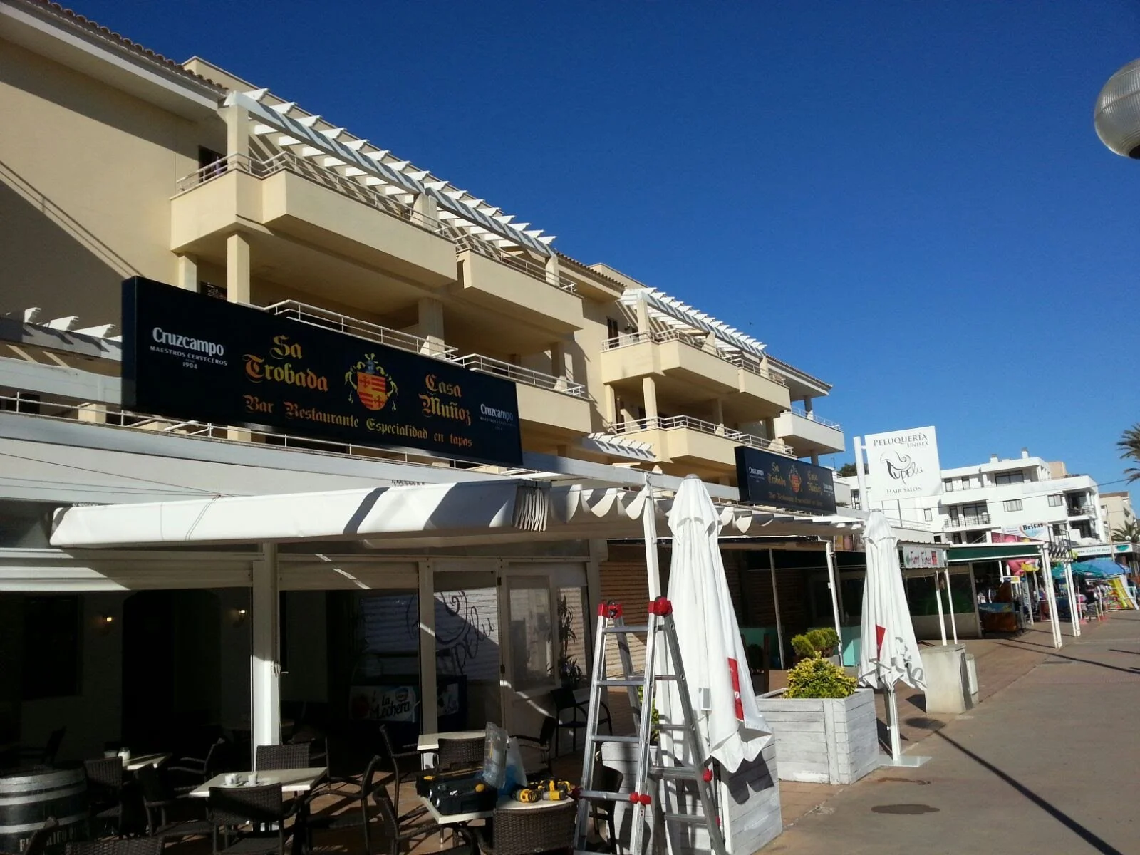 Vista de un edificio con varios restaurantes en la planta baja y apartamentos en los pisos superiores, en un día soleado con cielo despejado.