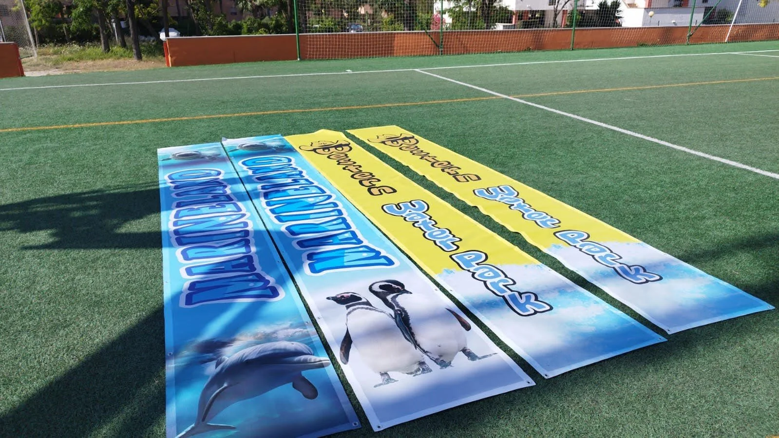 Tres lonas promocionales en una cancha de fútbol con imágenes de pingüinos, delfines y patos. La lonas están en colores azul y amarillo, con textos en inglés y español.