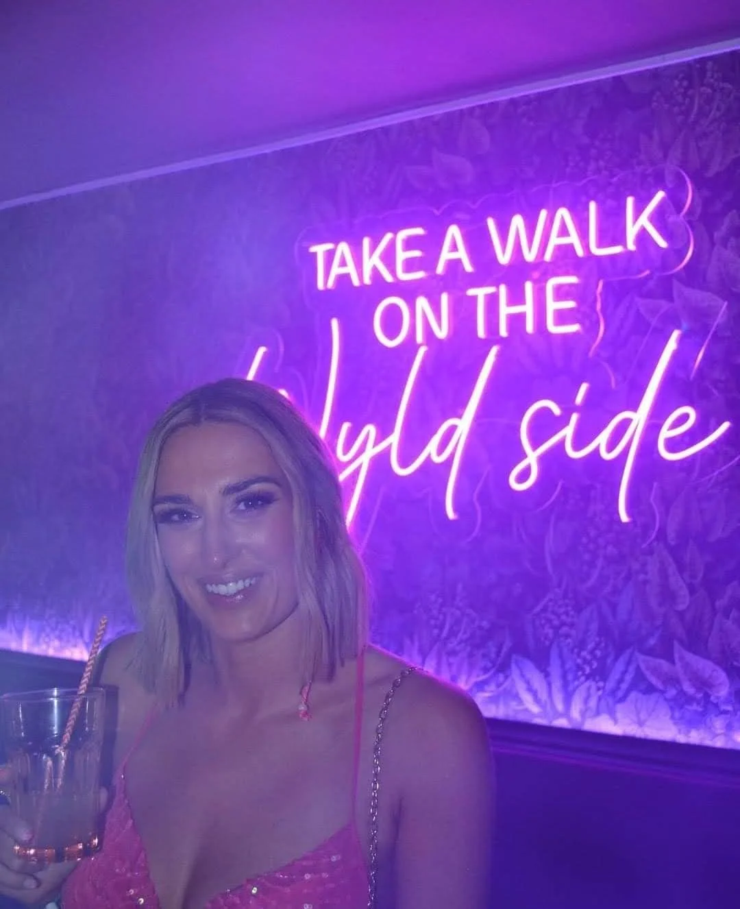 Mujer joven con blusa rosa en un lugar con una pared de fondo con cartel de neón rosa que dice 'Toma un paseo en el lado salvaje'.