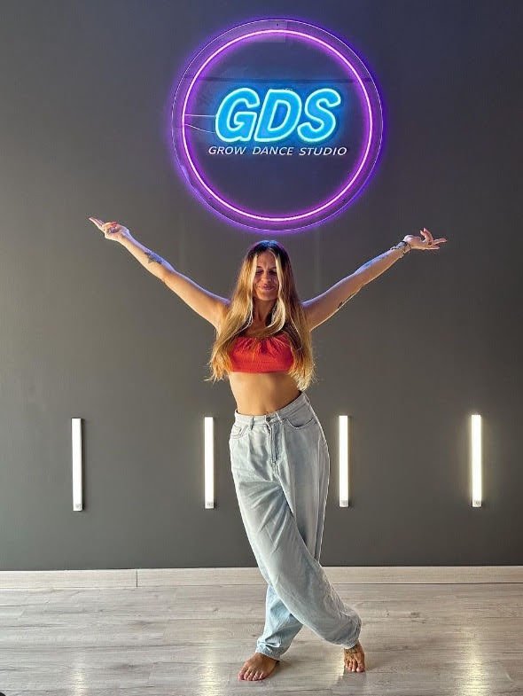 Joven bailando frente a un cartel luminoso que dice 'GDS Grow Dance Studio', con brazos extendidos y expresión de alegría, en un estudio de danza con paredes grises y decoracion minimalista.