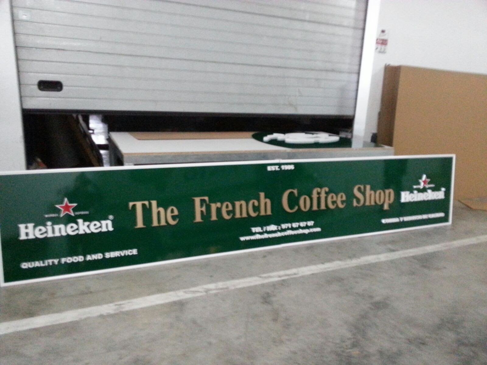 Cartel de un café llamado 'The French Coffee Shop' con logotipo de Heineken, ubicado en un estacionamiento con fondo de pared de garaje metálico y caja de cartón a la derecha.
