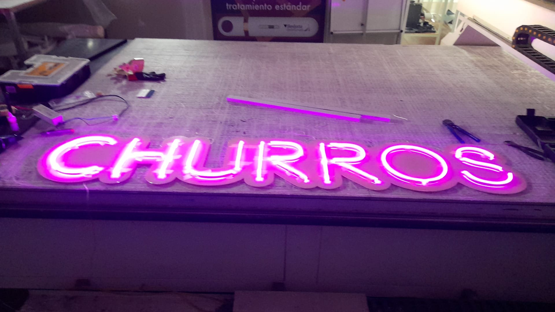 Letrero de neón rosa que dice 'CHURROS', en un ambiente de taller con herramientas y materiales de fabricación.