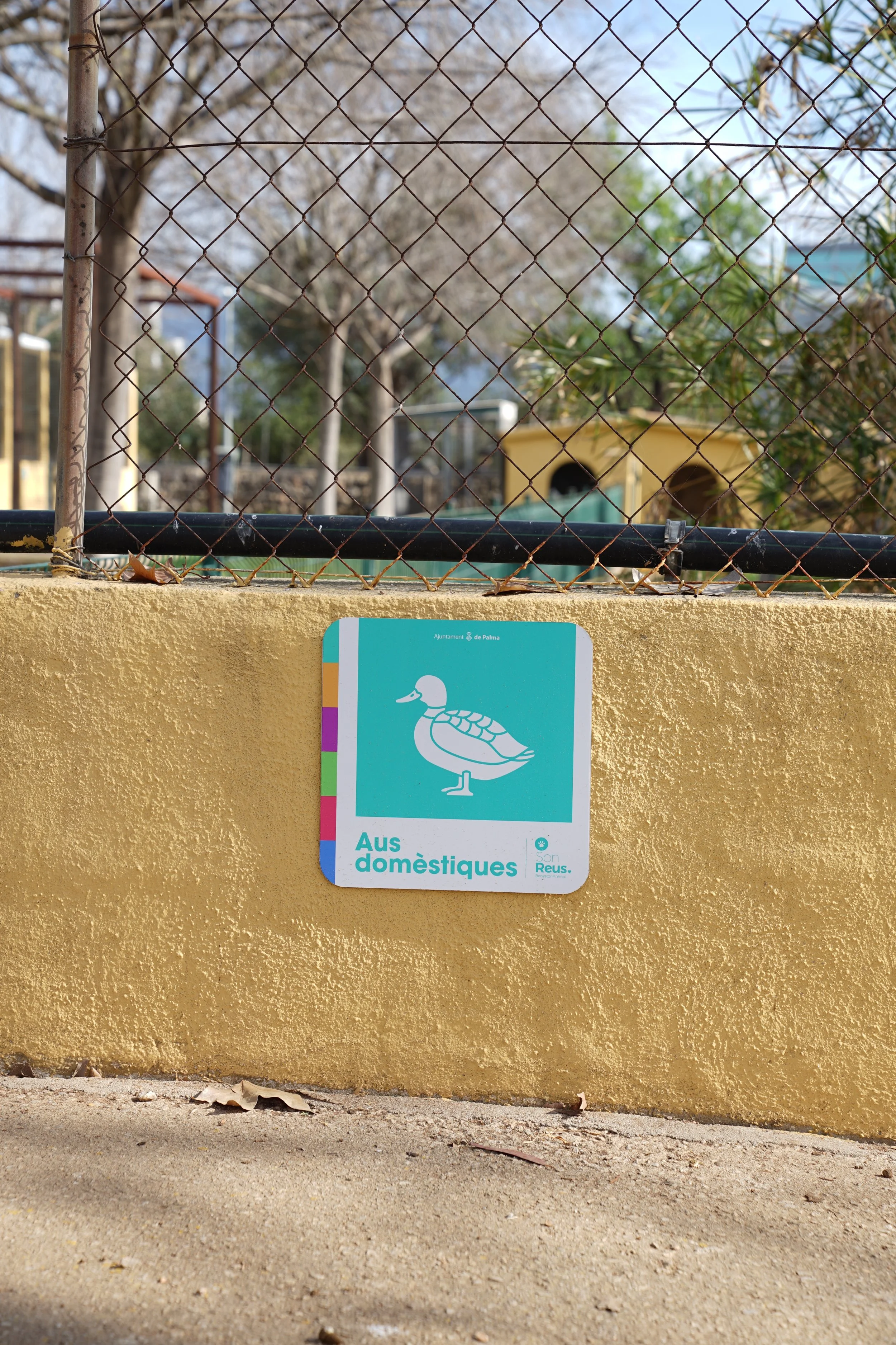 Señal en tono azul que indica área para aves domésticas, colocada en un muro amarillo cerca de una cerca de alambre, con árboles y edificios en el fondo.