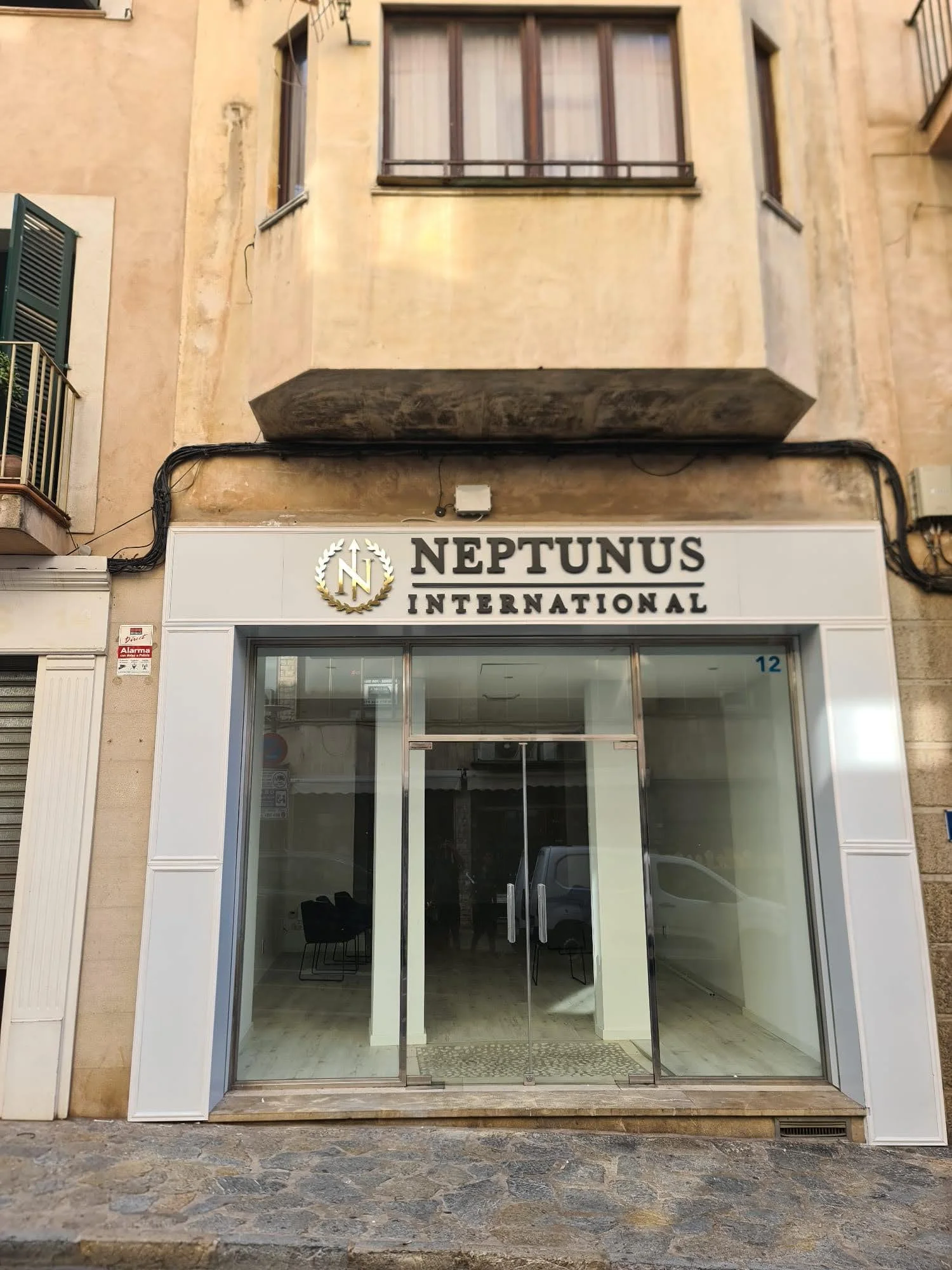Fachada de un edificio con un letrero que dice 'NEPTUNUS INTERNATIONAL', con ventanas y puertas de vidrio y un piso de piedra.