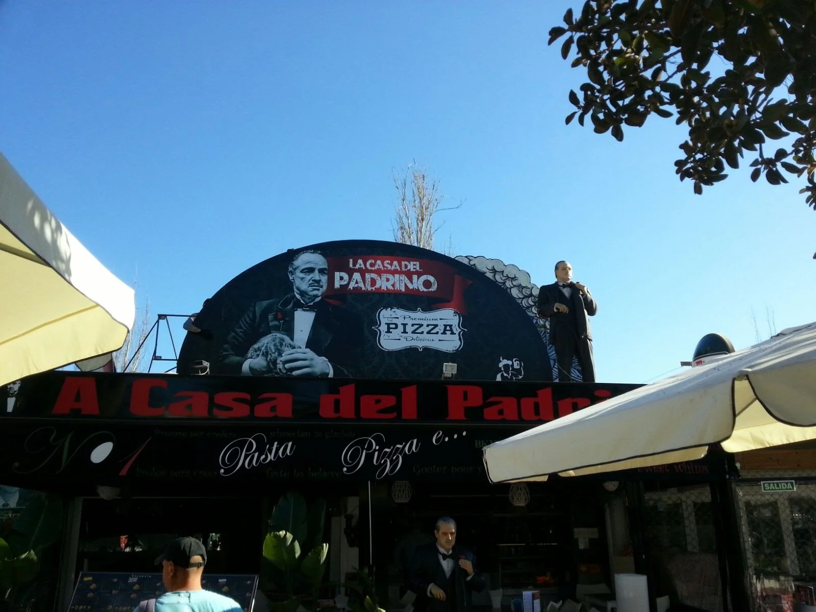 Cartel del restaurante 'La Casa del Padrino' con la imagen de un hombre vestido en esmoquin y sosteniendo un plato de pizza, en un fondo azul del cielo y árboles.