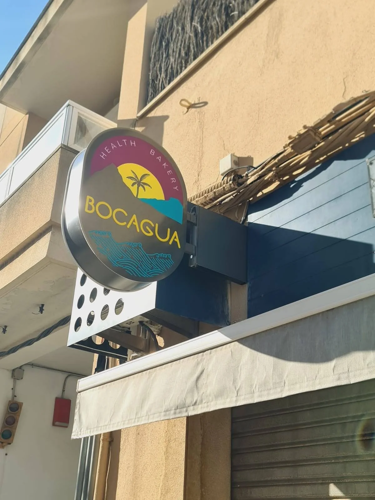 Cartel de negocio con el nombre 'Bocagua', que muestra una palmera, agua y un sol en un fondo de montañas, y la frase 'Health Bakery'.