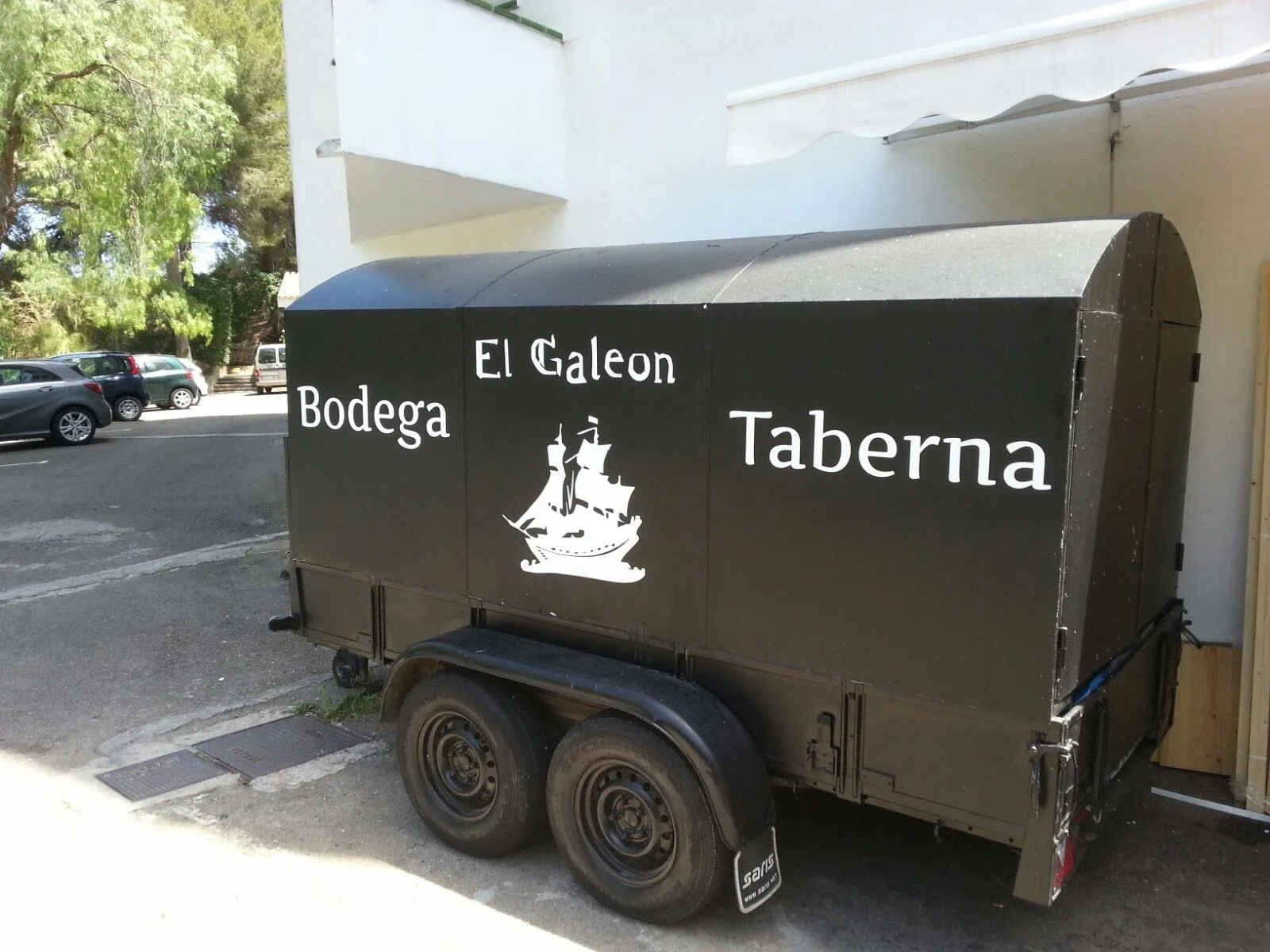 Un remolque negro con letreros blancos que dice 'Bodega El Galeón Taberna' y tiene una imagen de un galeón en el centro, estacionado junto a un edificio y en un fondo de árboles.