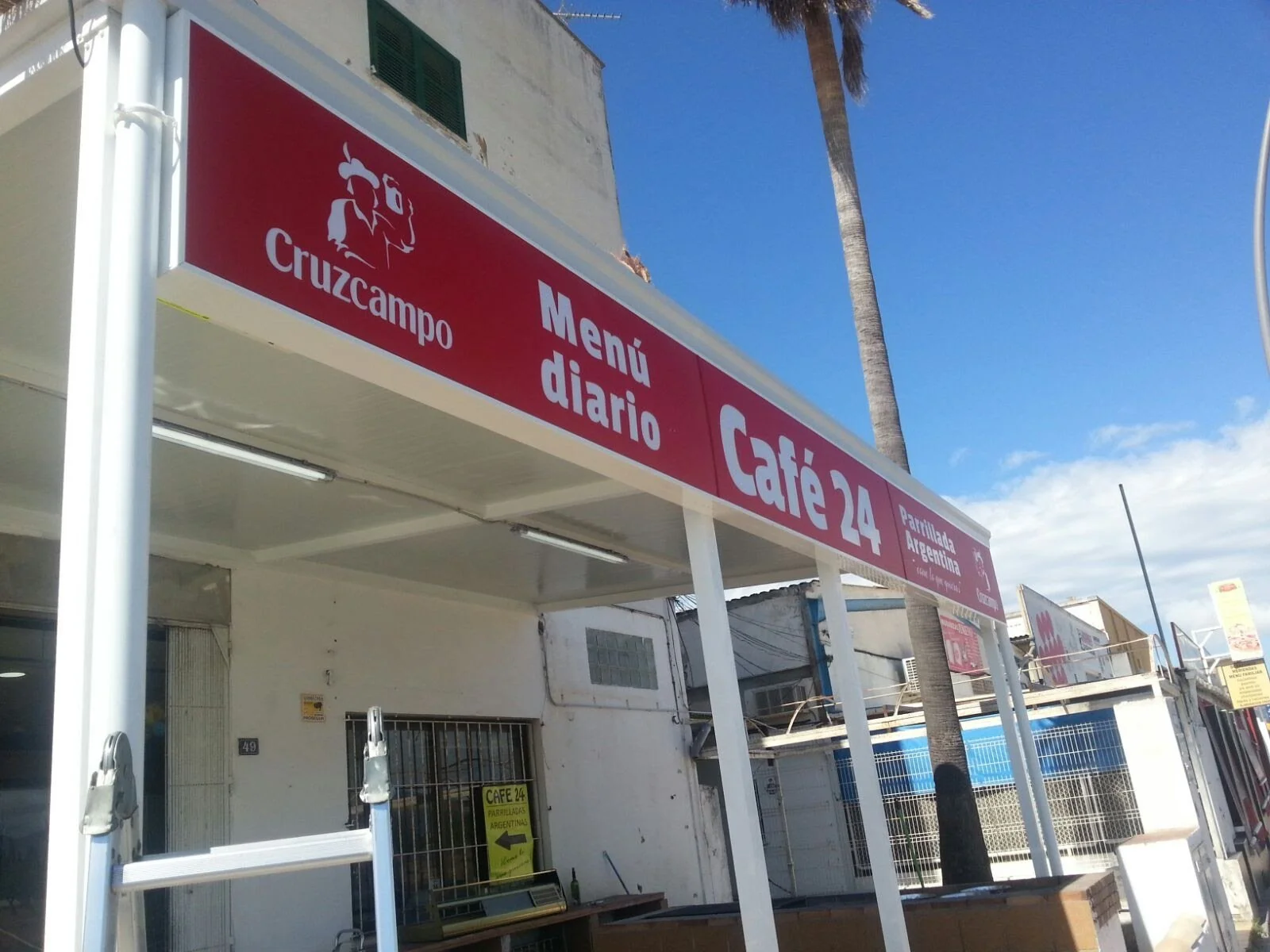 Cartel de un restaurante llamado Café 24 con señalización de menú diario, en un ambiente exterior con palmera y cielo claro.