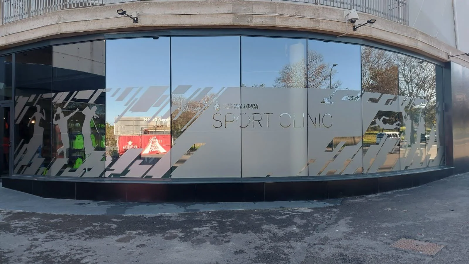Fachada de una clínica de deportes con ventanas de cristal decoradas con siluetas de deportistas y el texto 'Clinica Deportiva Sport Clinic'.