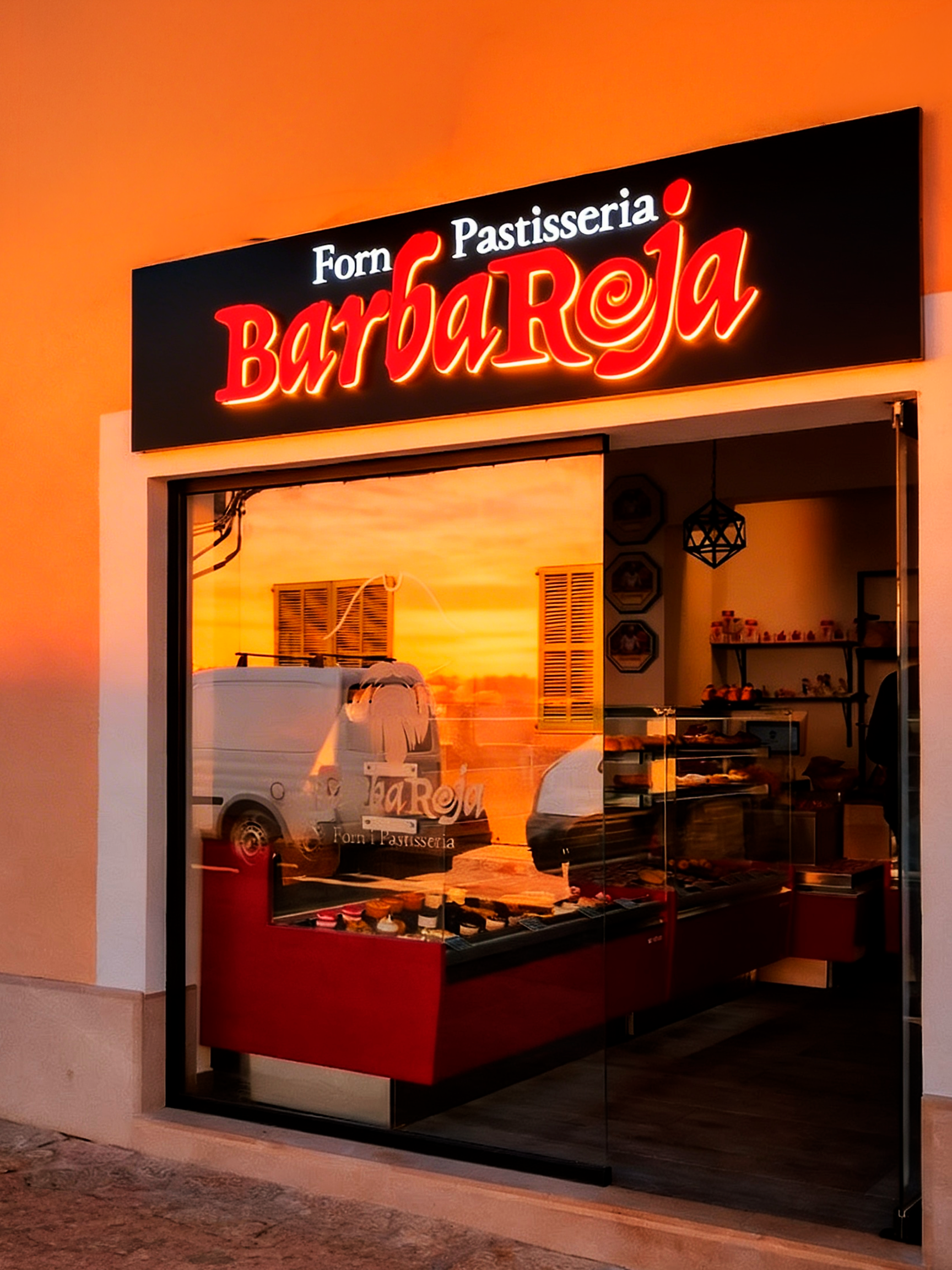 Fachada de panadería llamada 'BarbaRoja', con letrero luminoso en letras rojas y blancas y ventana que refleja una camioneta y la puesta del sol