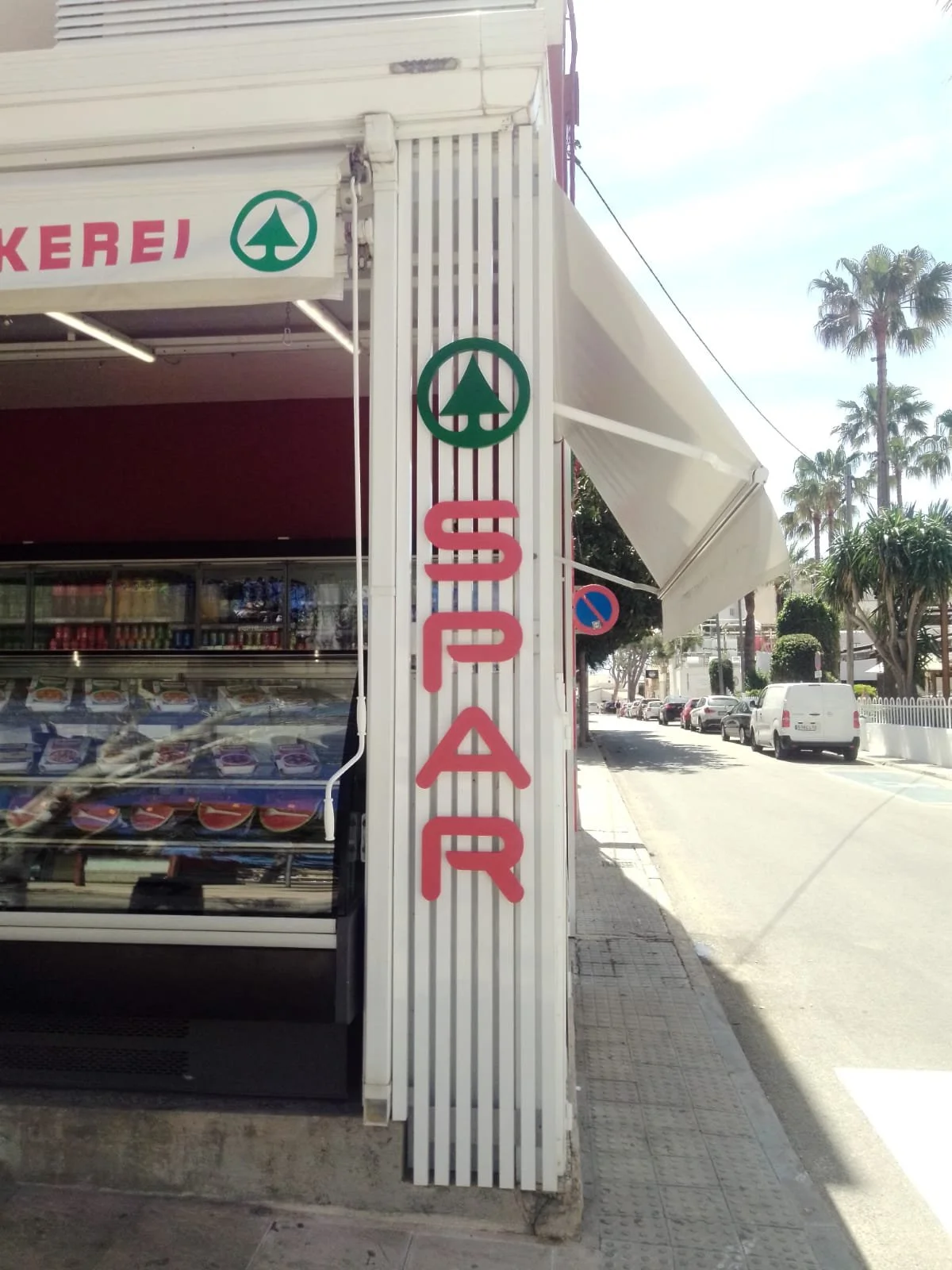 Entrada a una tienda con letrero de 'SEAL' en letras rojas y un símbolo verde de árbol. La tienda tiene una estructura de metal blanca y enfrente una calle con autos y palmeras.