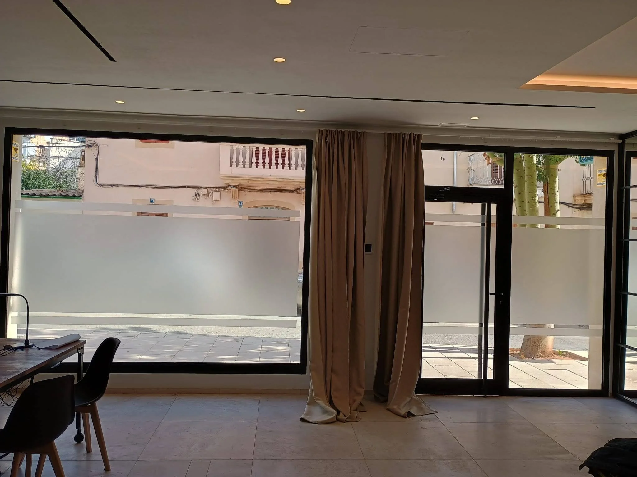 Interior de una sala con ventanas grandes y cortinas beige. Afuera se ven árboles, una banqueta y edificios.